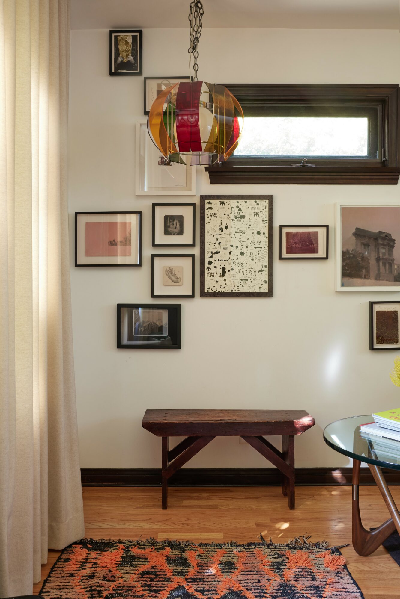 Interior wall with framed artwork, a wooden bench, and a window, with a colorful hanging lamp above.