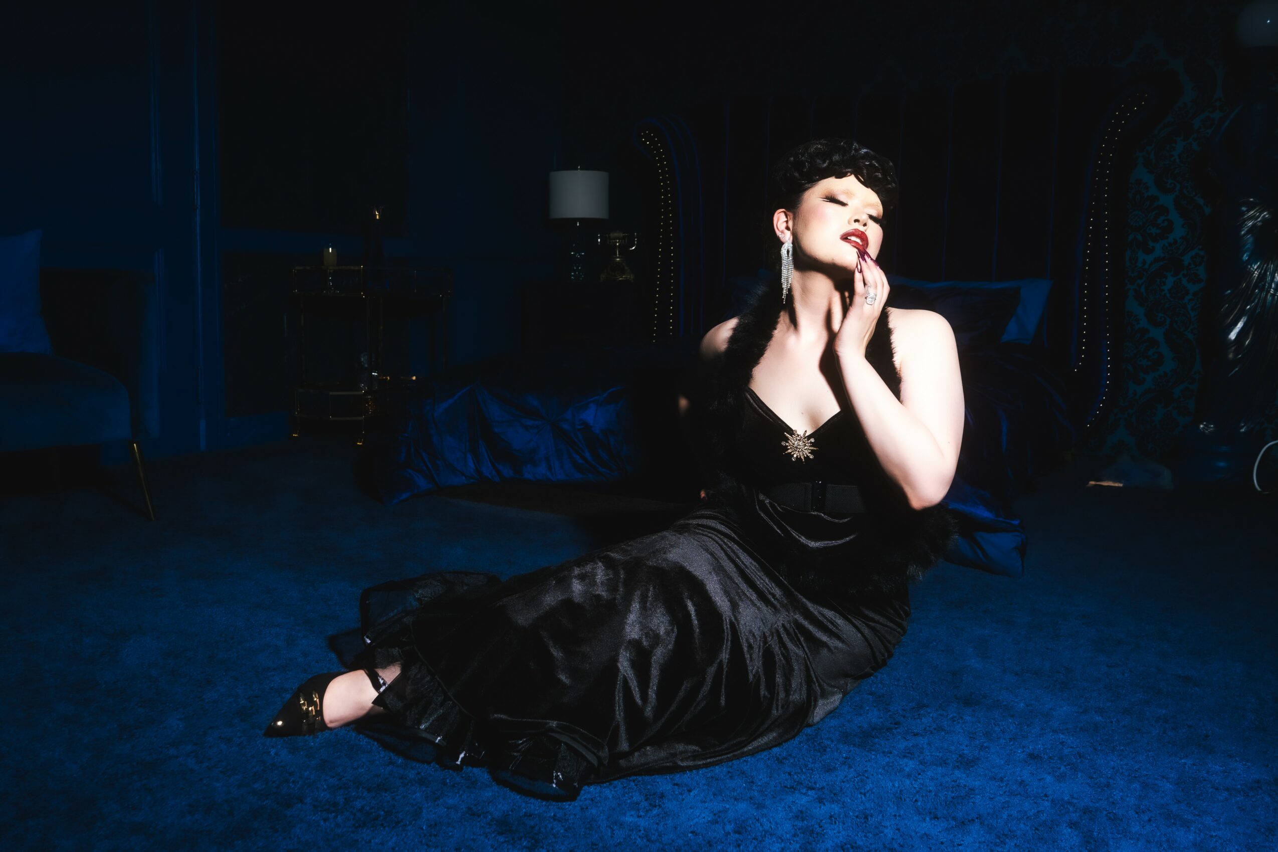Woman in black dress sitting on blue carpet with hands near face, dimly lit room background, elegant setting.