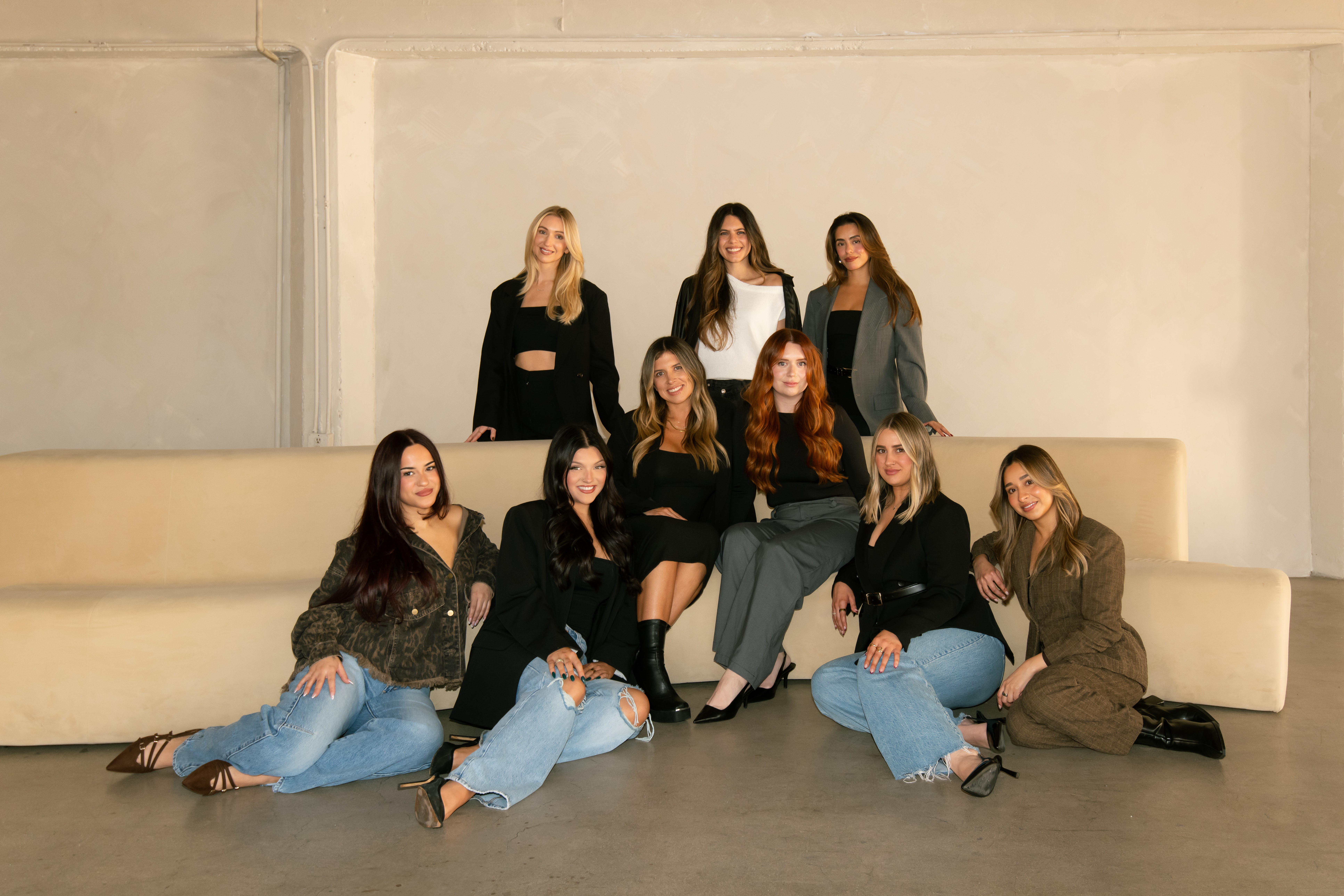 Group of nine women sitting and standing on a beige sofa in a plain room, posing for a photo.