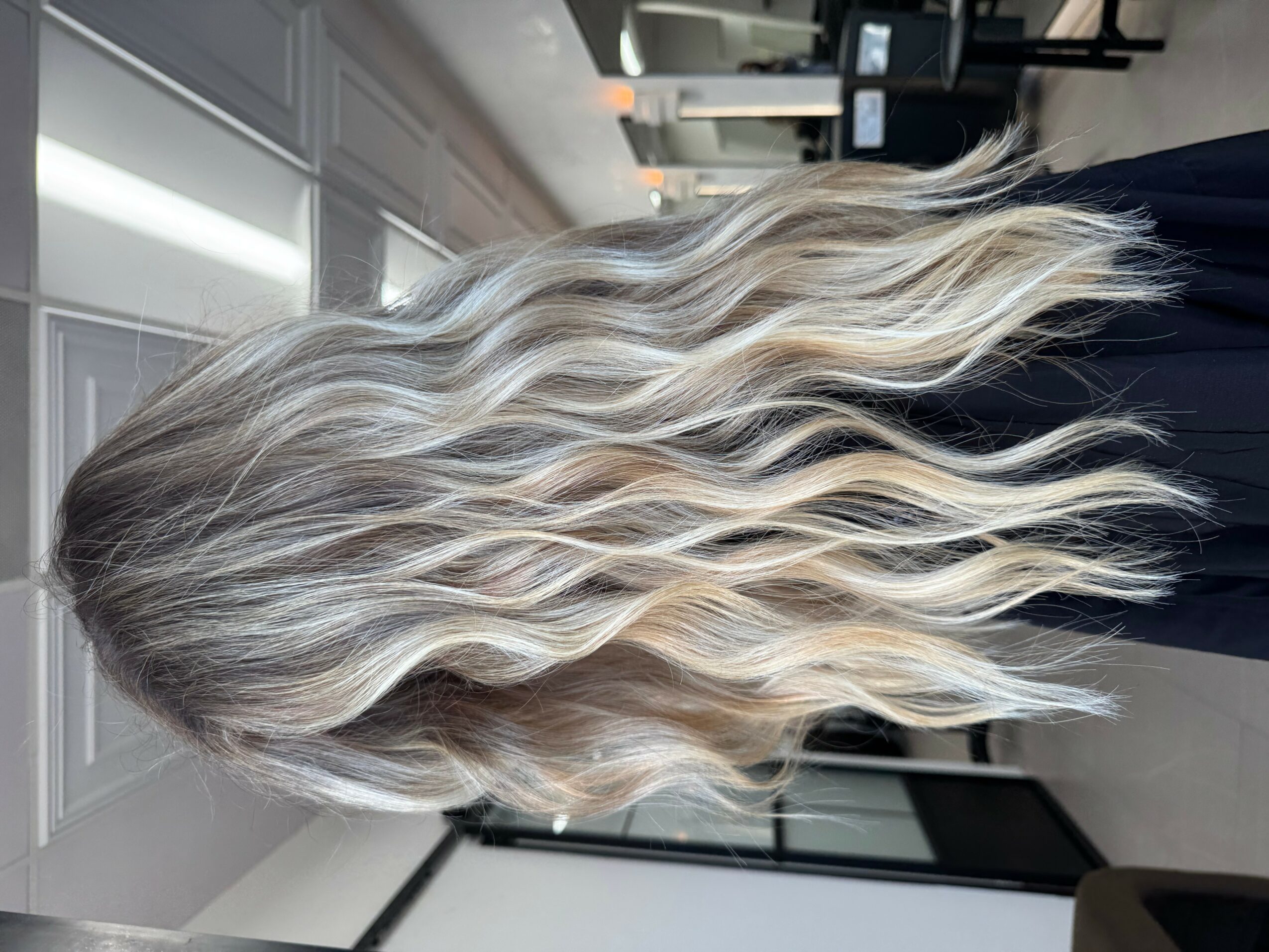 Person with long, wavy, blonde hair standing indoors, facing away from the camera.