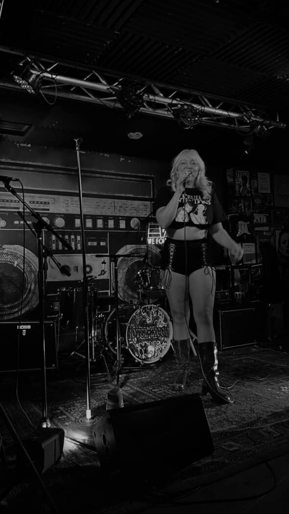 Female singer with curly hair performs on stage with drums and equipment behind her, in a dark setting.