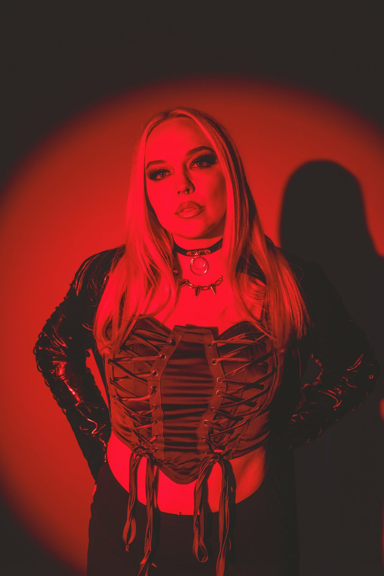 Woman with long hair, wearing a choker and a corset-style top, standing with hands on hips, illuminated by red light
