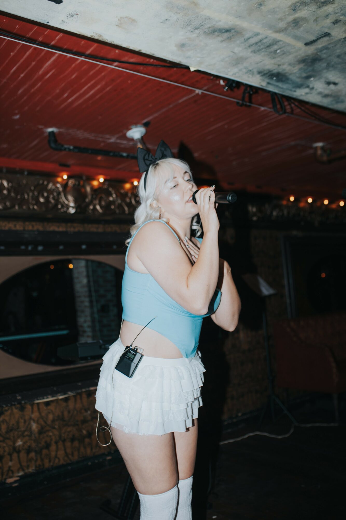 Young woman with blonde hair singing into a microphone in a dimly lit venue, wearing a light blue top and white shorts.