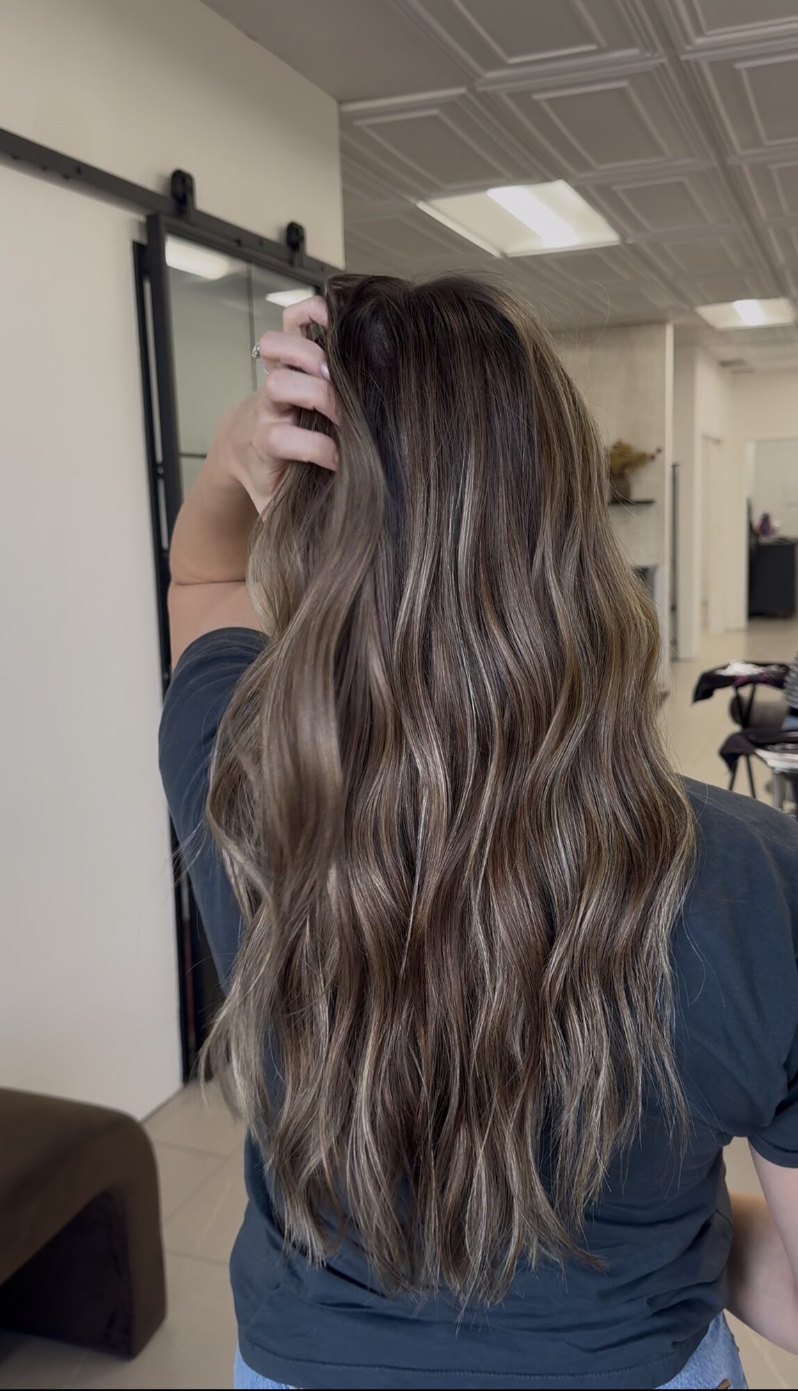 Person with long, wavy hair standing indoors, hand touching hair, facing away from camera.