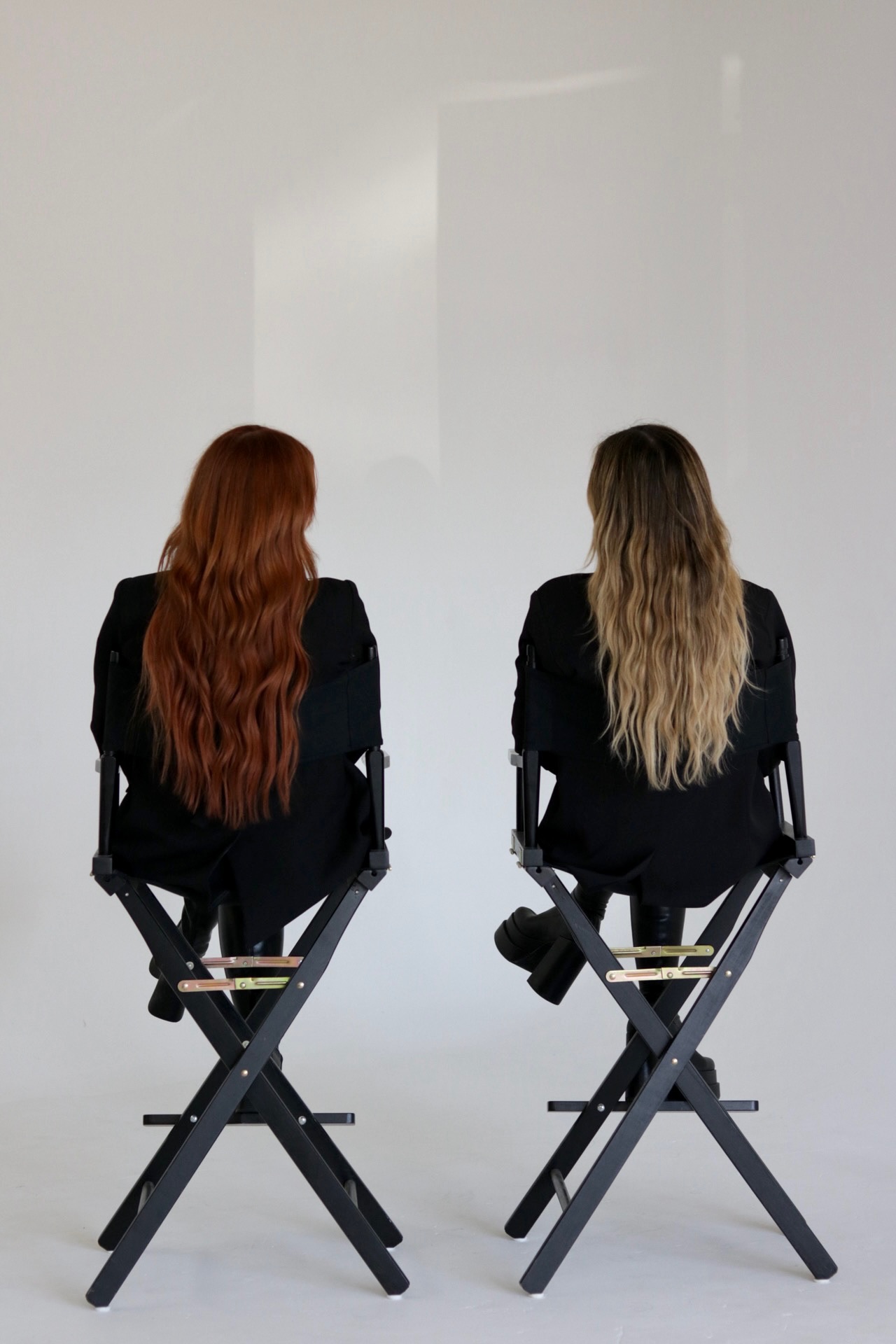 Two women with long hair sit on chairs facing away, against a plain light background.