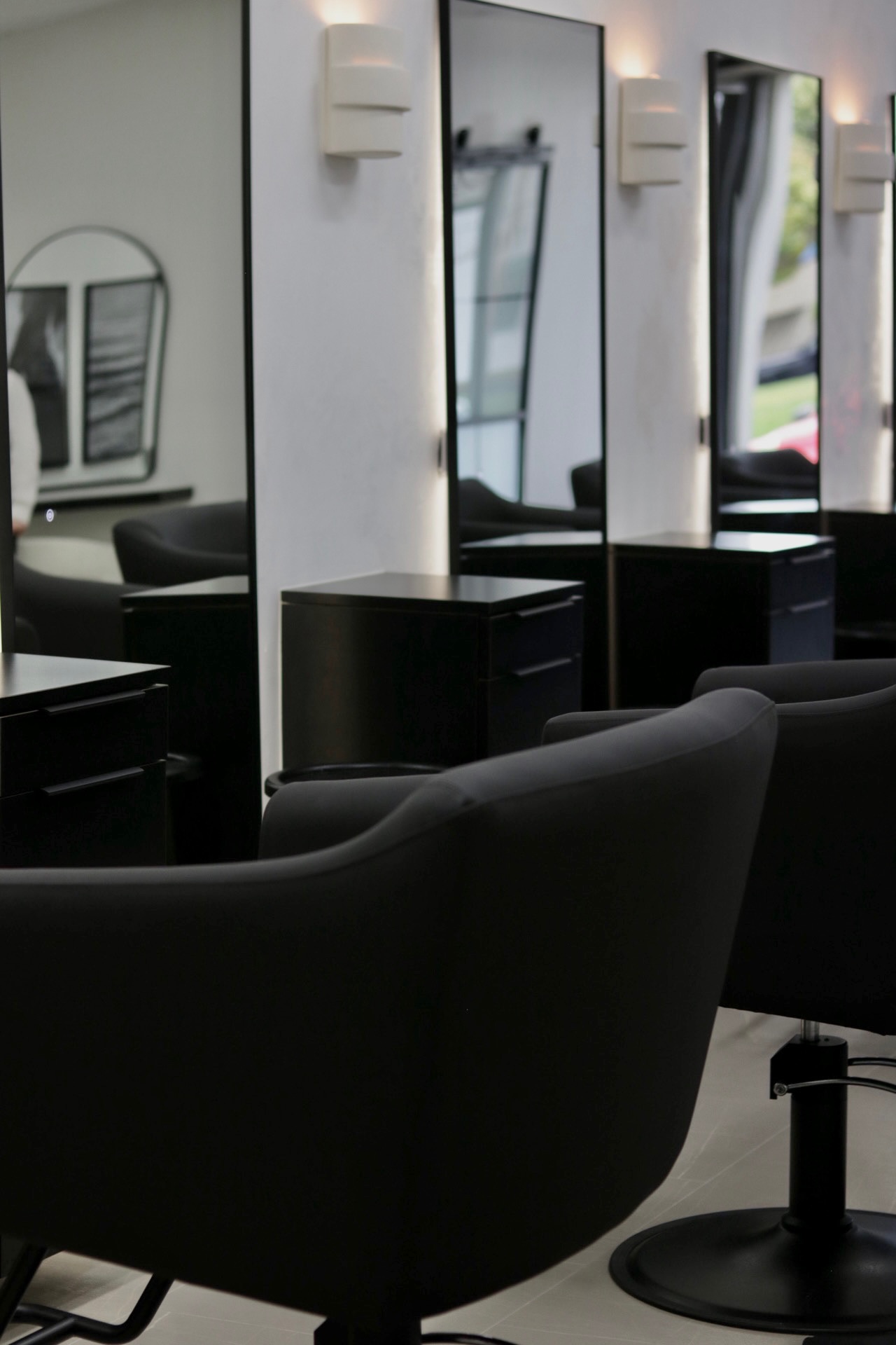 Salon with mirrors, chairs, and styling stations, illuminated by wall lights, with a window reflecting outside view.