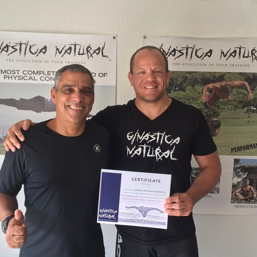 Two men smiling, one holding a certificate, standing in front of posters with text and images related to physical training.
