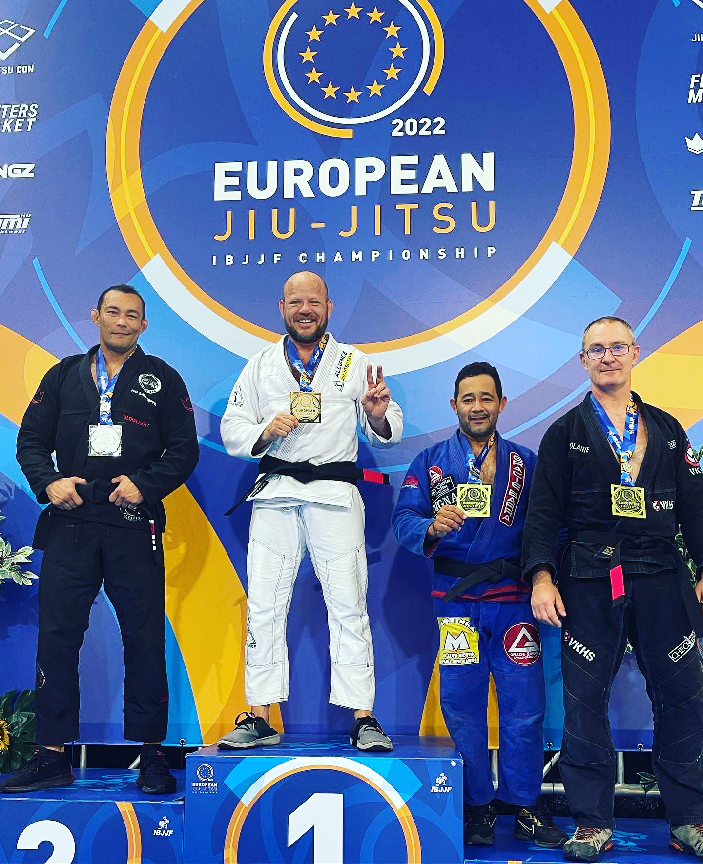 Four men on a podium at the European Jiu-Jitsu Championship, with medals and a large banner behind them, celebrating victory.