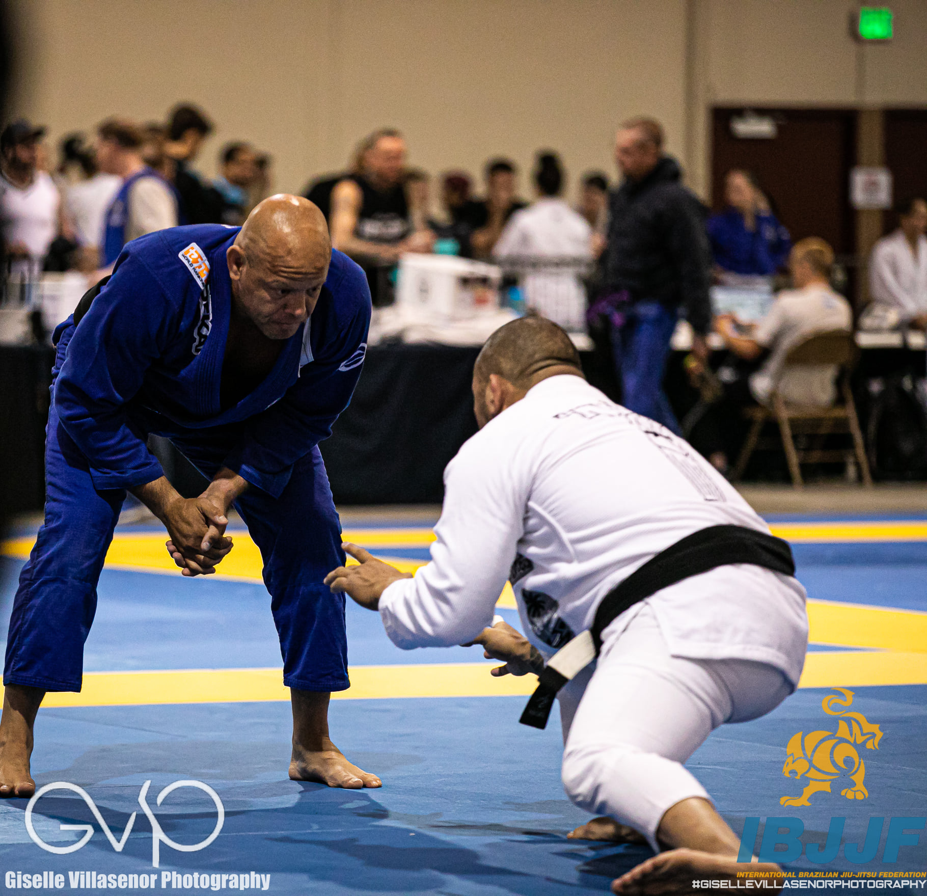 Two martial artists in gis practicing on a mat, with a group of people in the background at an indoor event.