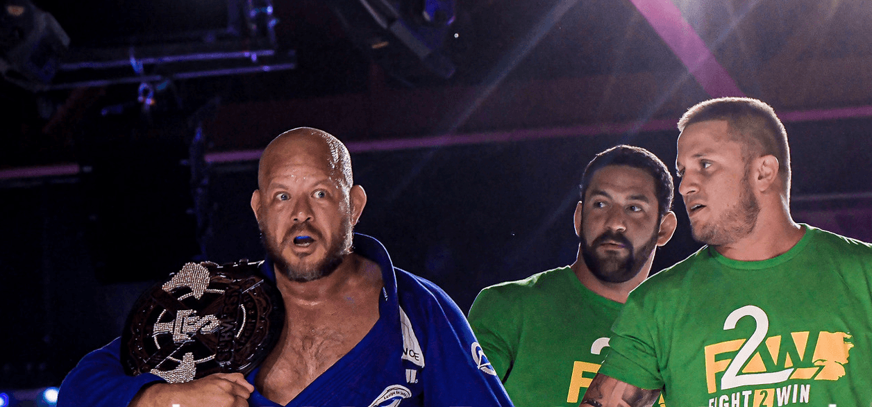 Three men standing together, two wearing green shirts and one in a blue jacket, in a dimly lit setting.