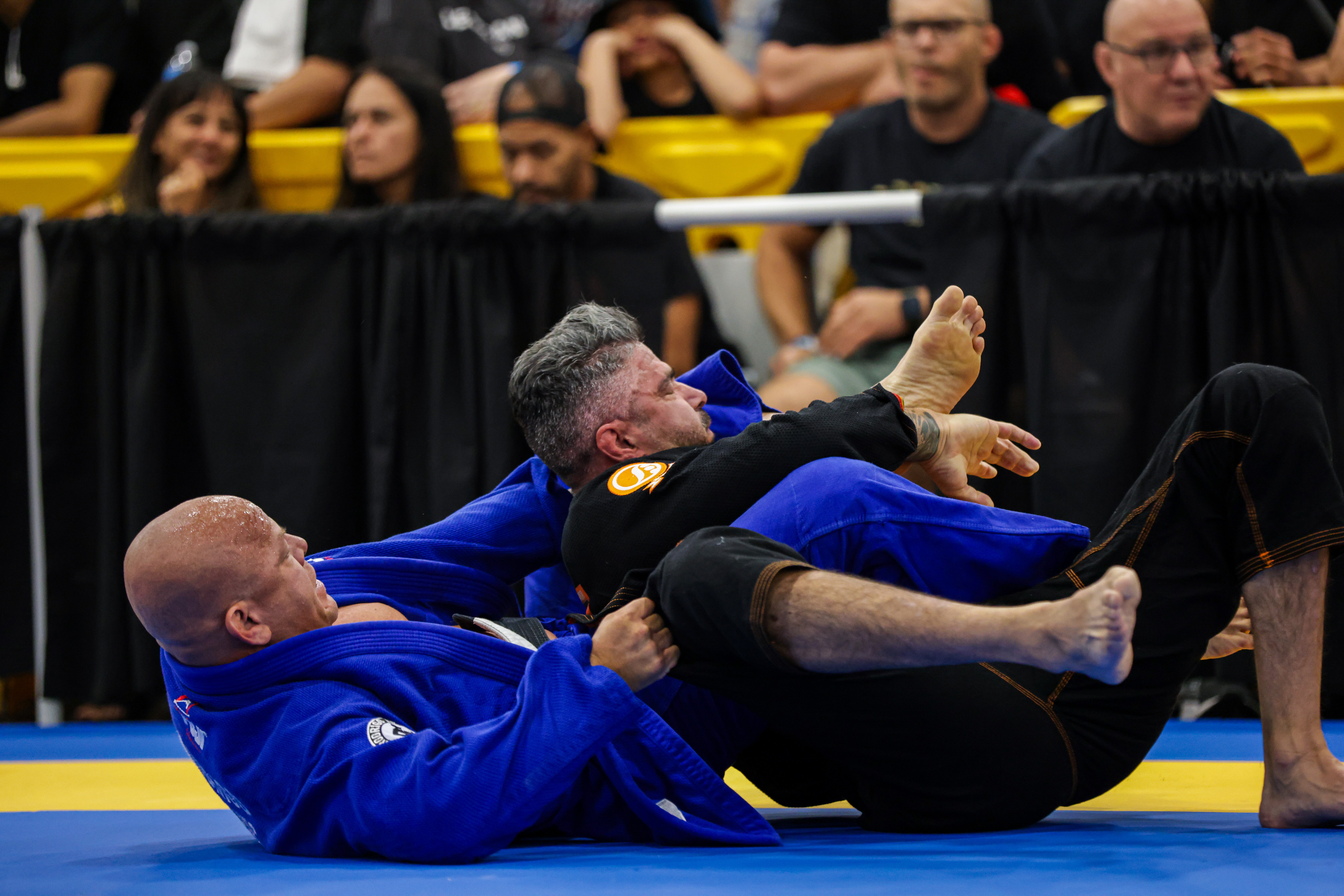 Two people in Brazilian Jiu-Jitsu gis grappling on a mat, with spectators watching in the background.