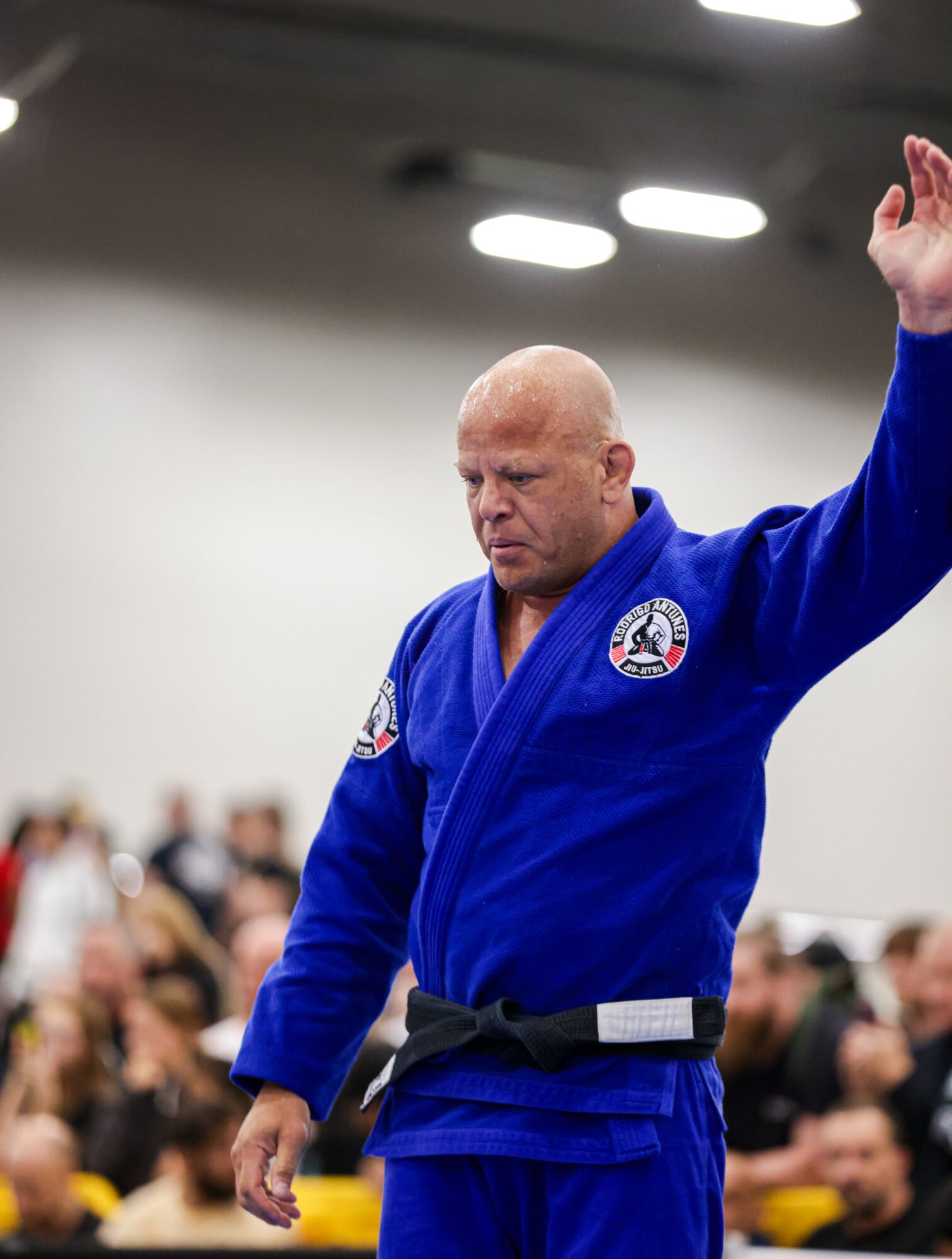 Bald man in blue martial arts uniform with black belt raising his right hand, in a gym with an audience.