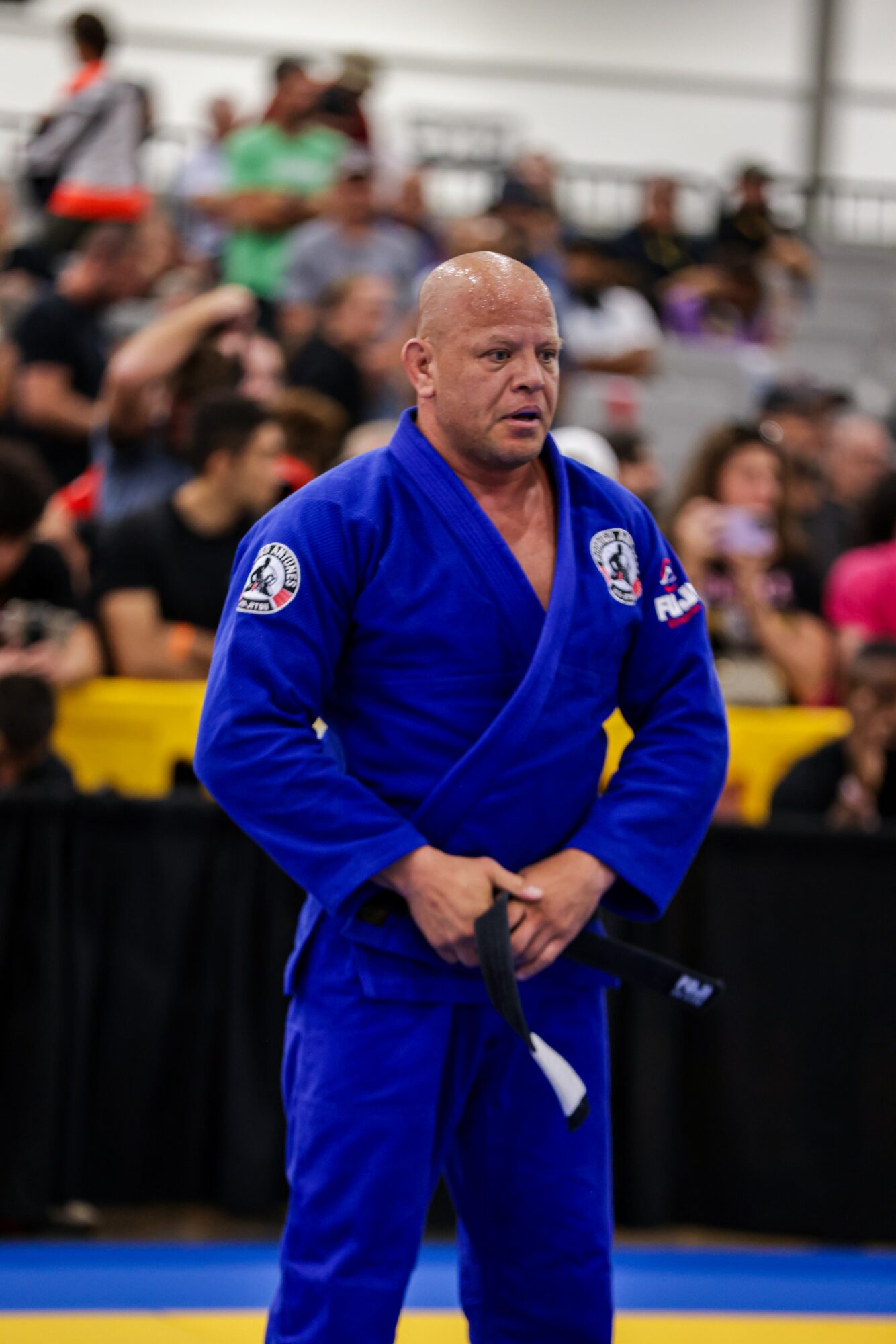 Martial artist in blue gi holding a black belt, standing in front of a crowd, indoors, with a focused expression.
