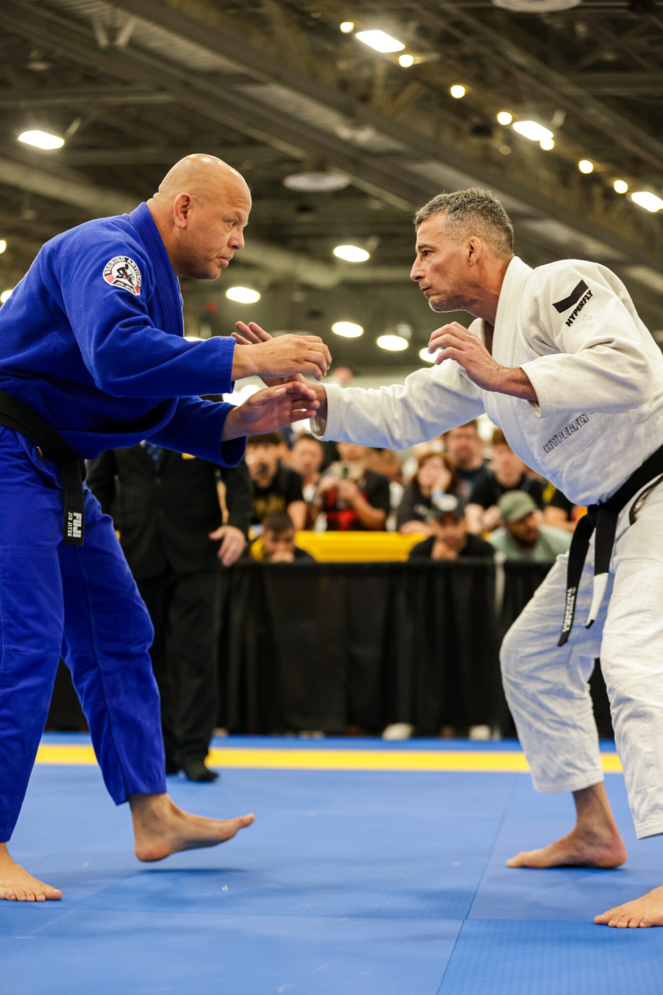 Two martial artists in gi uniforms shake hands on a blue mat, with a crowd and tables in the background.