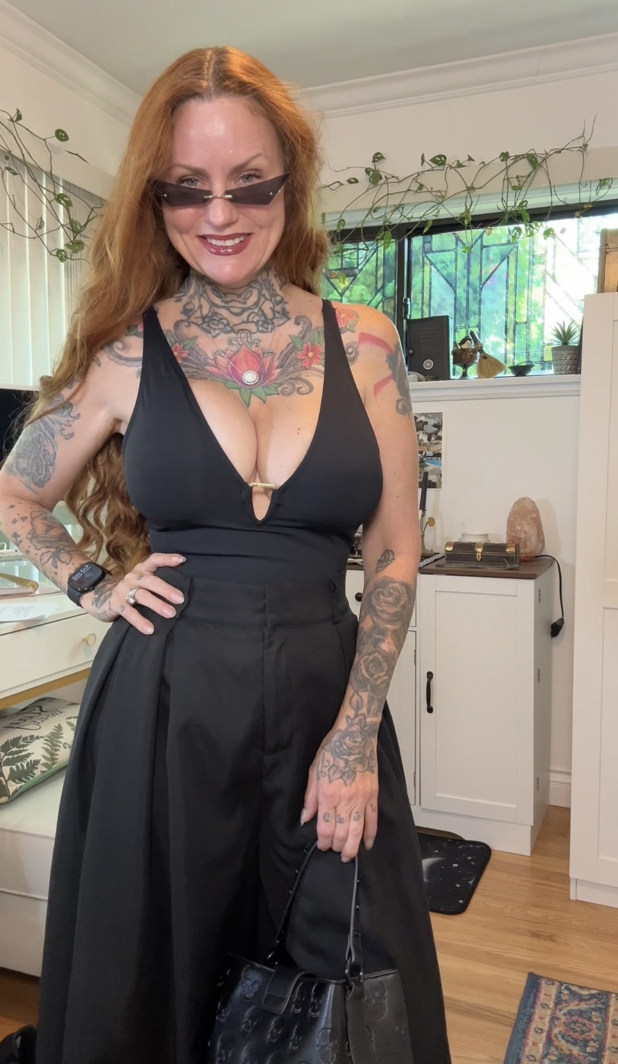 Woman with long red hair, tattoos, wearing sunglasses, black dress, standing indoors with a window and greenery behind her.