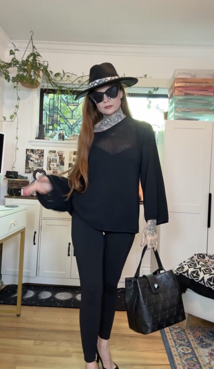 Woman in black outfit with hat and sunglasses holding a black handbag, standing indoors near a window and white furniture.