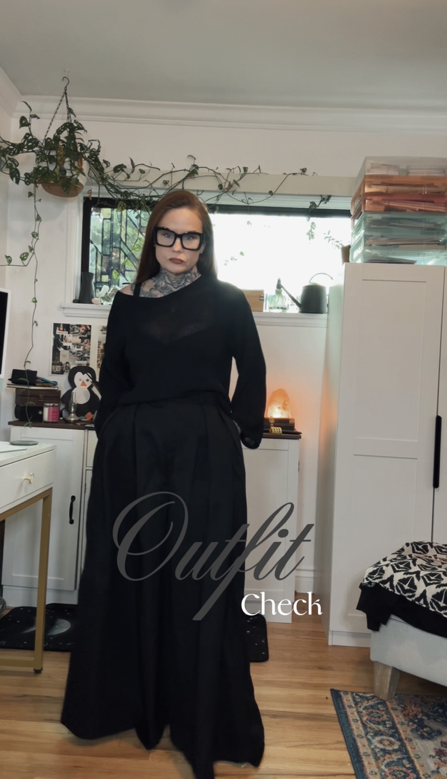 Woman in black outfit with glasses standing indoors near a window and white furniture.