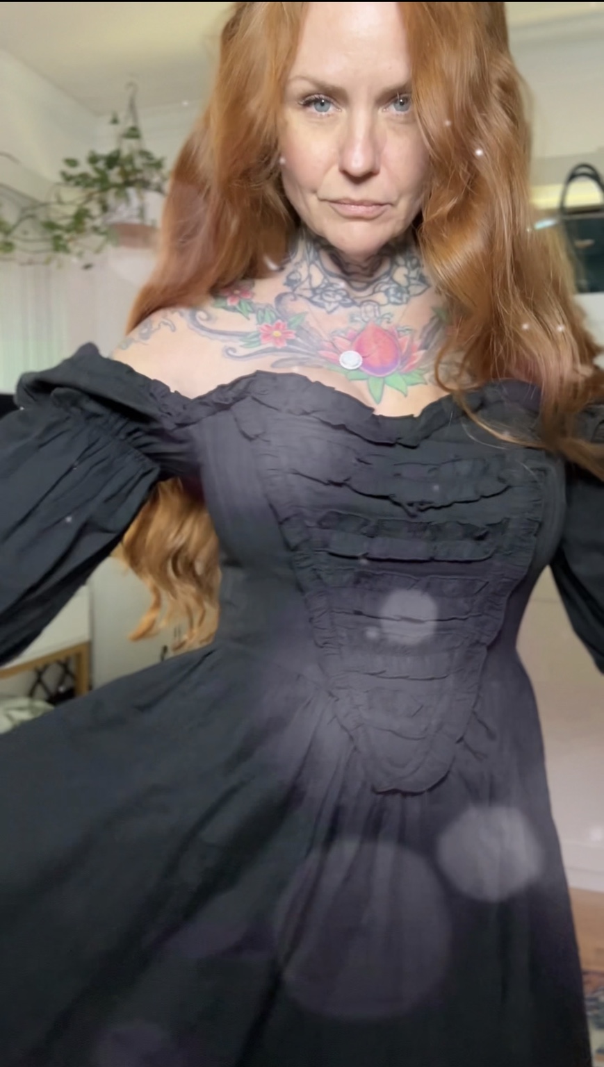 Woman with long red hair wearing a black off-shoulder dress, showing colorful tattoos on her chest and neck.