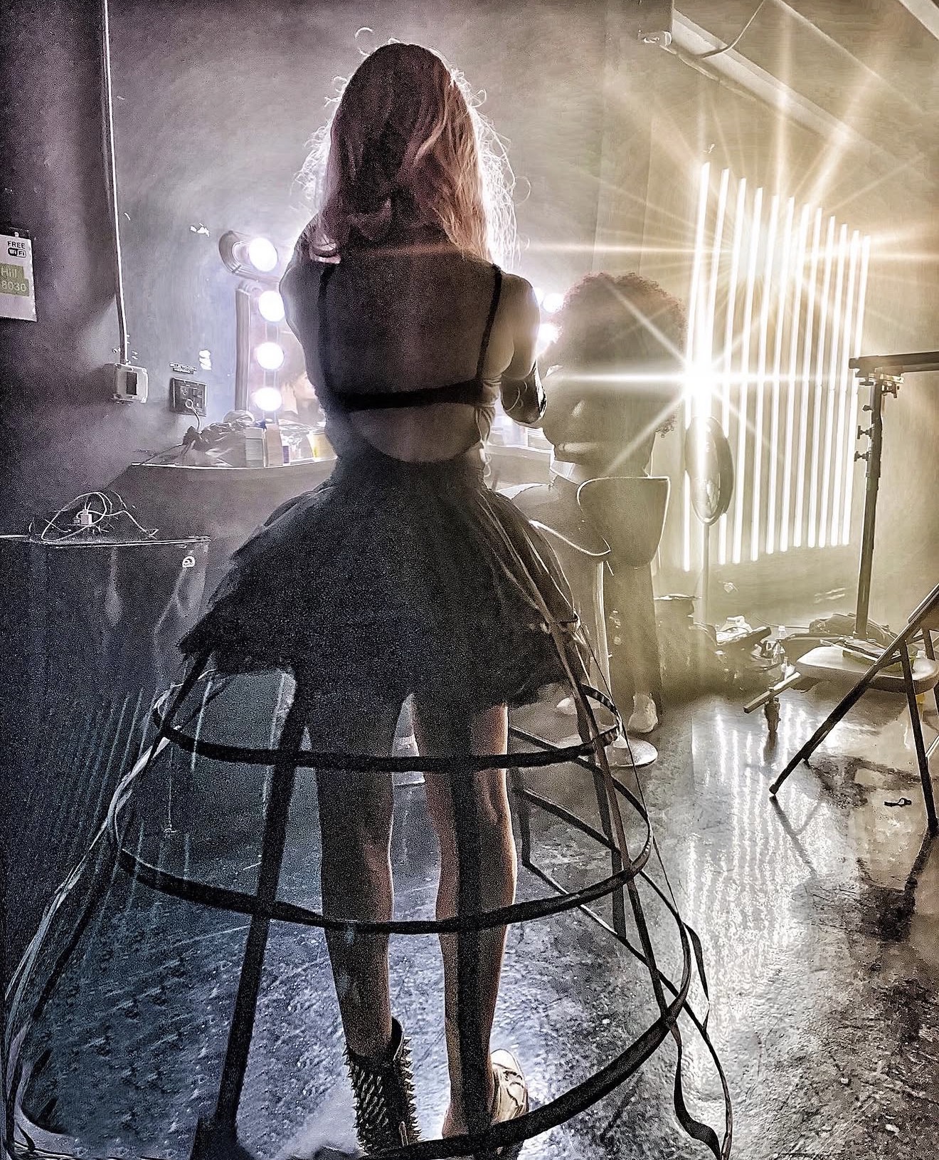 Person with long hair wearing a black dress and fishnet stockings, standing in a room with bright lights and equipment.