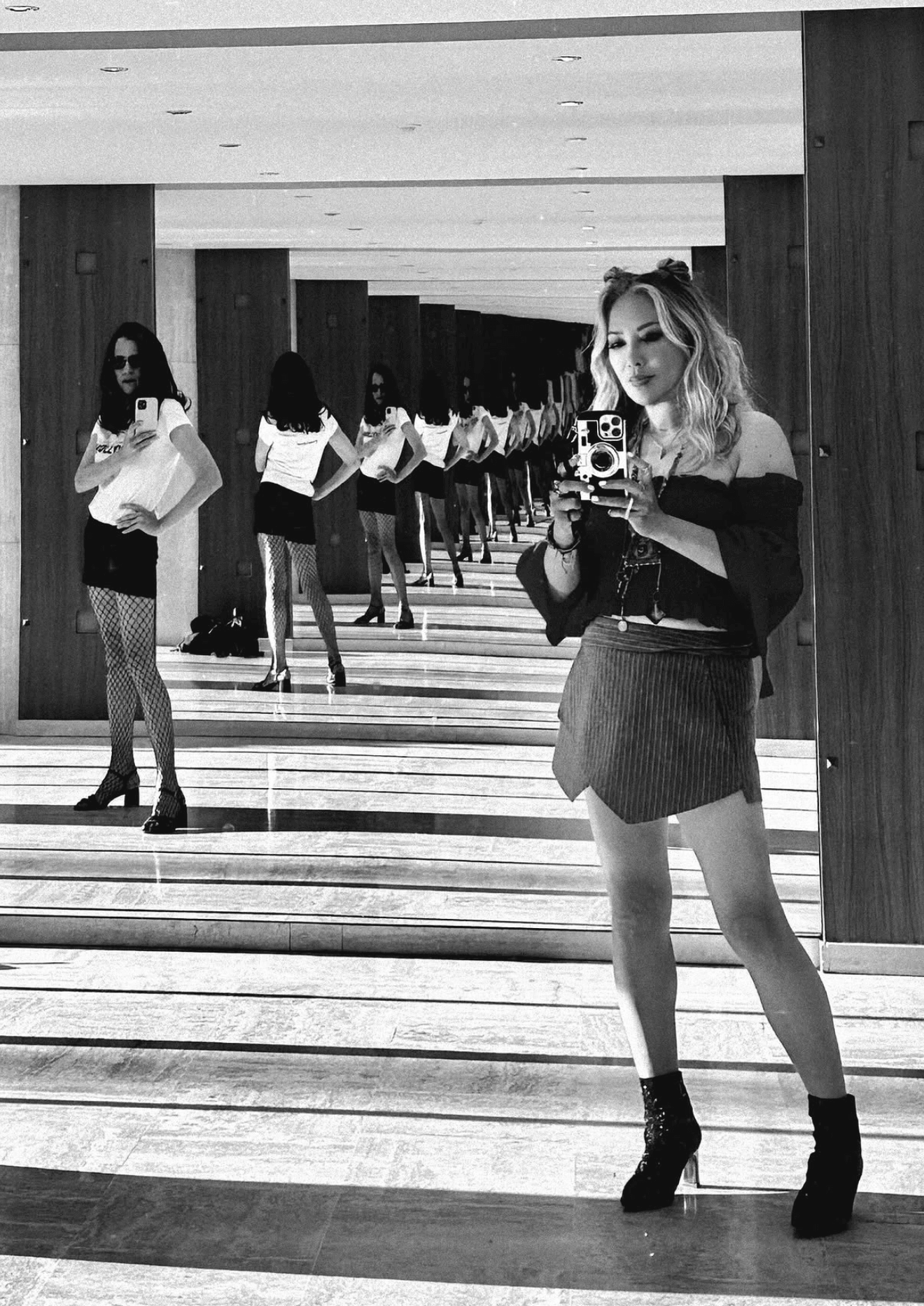 A woman taking a mirror selfie in a room with multiple reflections of herself, wearing a skirt, boots, and a top.
