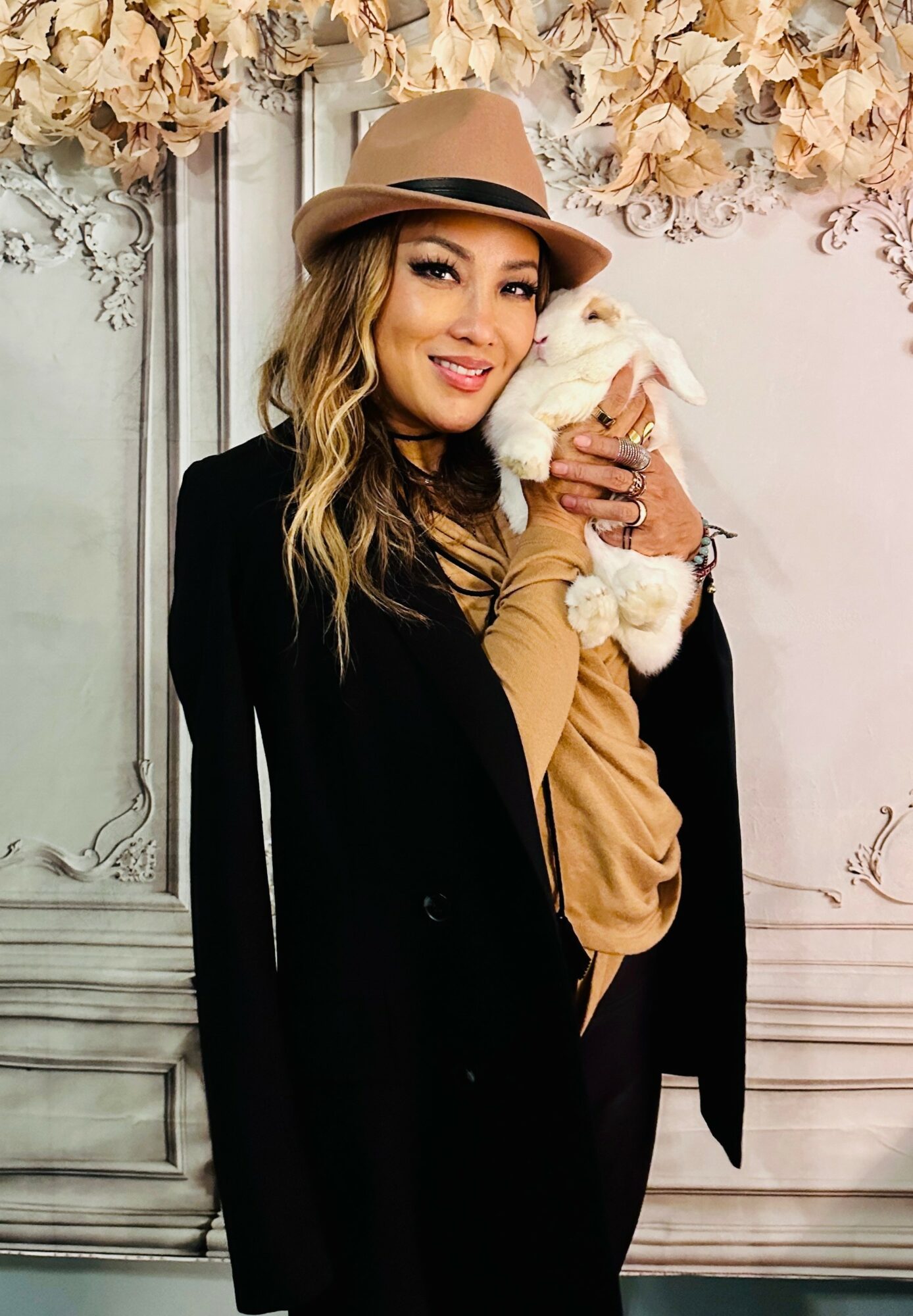 Woman with long hair wearing a hat and black blazer holding a small white dog, standing in front of a floral backdrop.