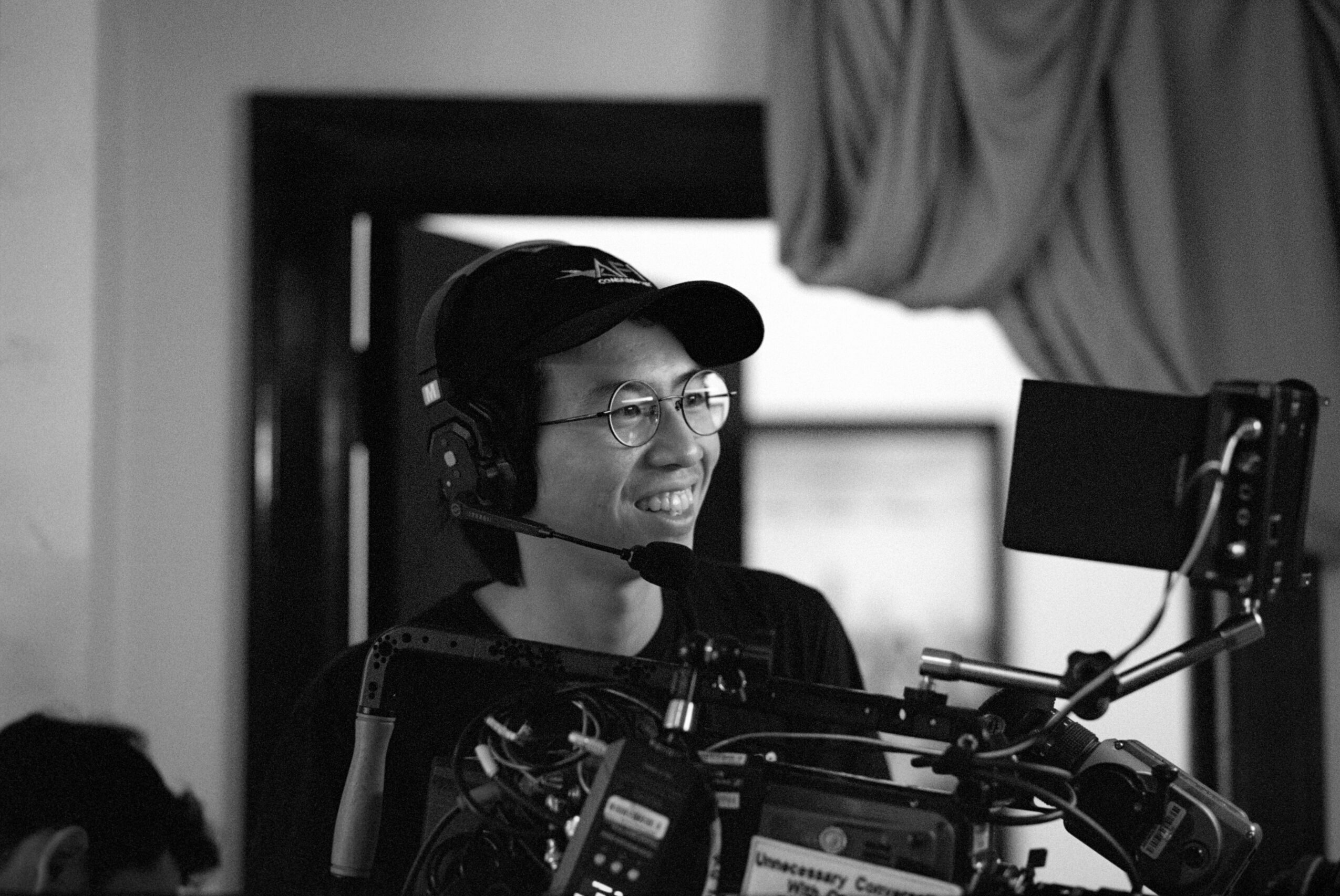 Person wearing headphones and glasses, smiling, operating camera equipment in a studio setting.