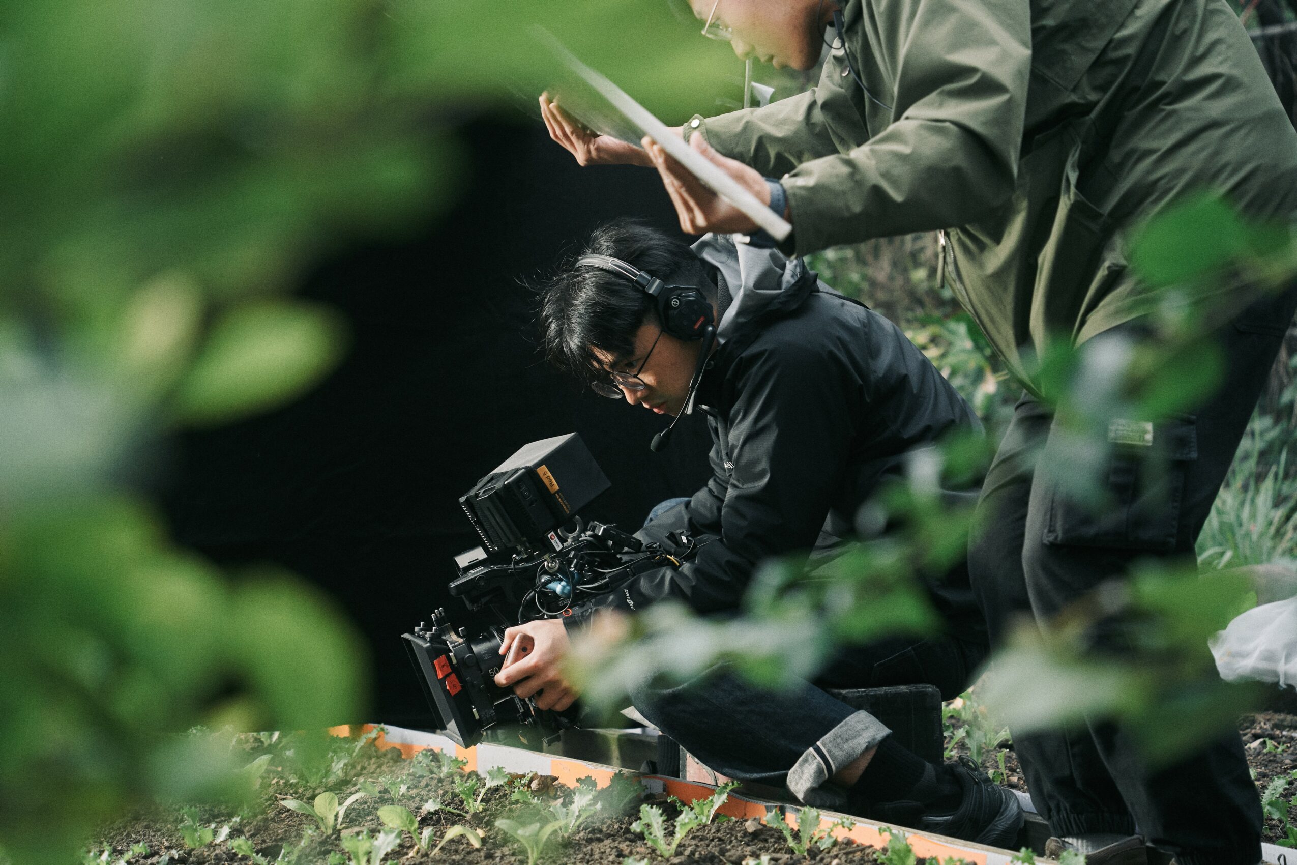 Two people working outdoors with camera equipment, surrounded by green foliage, one person adjusting the camera while the other reads a notebook.