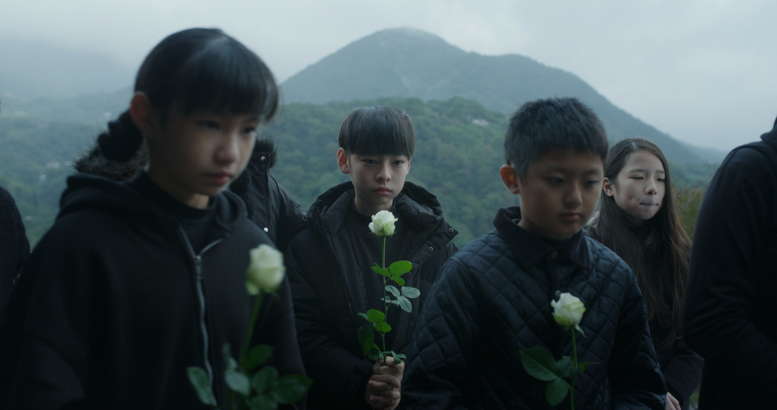 Group of people holding white roses outdoors with mountains in the background, overcast sky, somber mood.