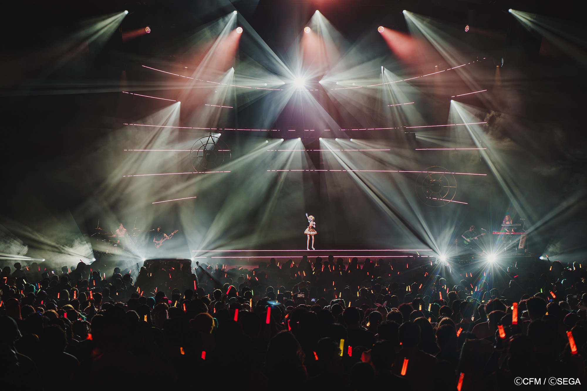 Stage with performer and colorful light beams, audience holding glow sticks, dark background, concert atmosphere.