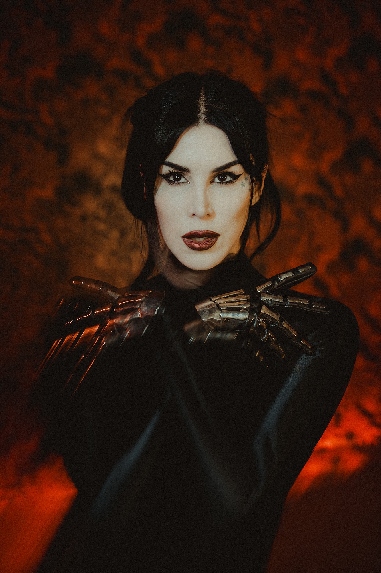Woman with dark hair and makeup, wearing black gloves, against a fiery orange background.