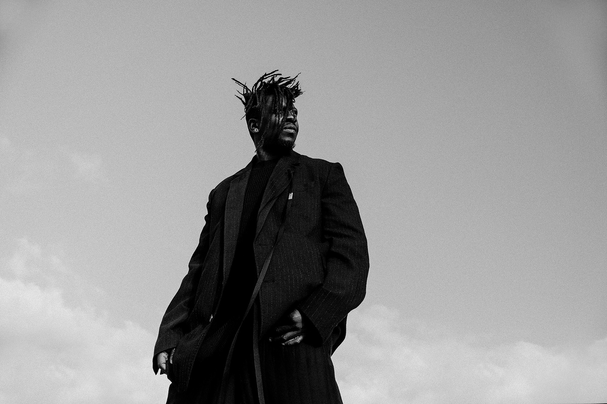 Person with dreadlocks wearing a dark coat stands outdoors against a cloudy sky.
