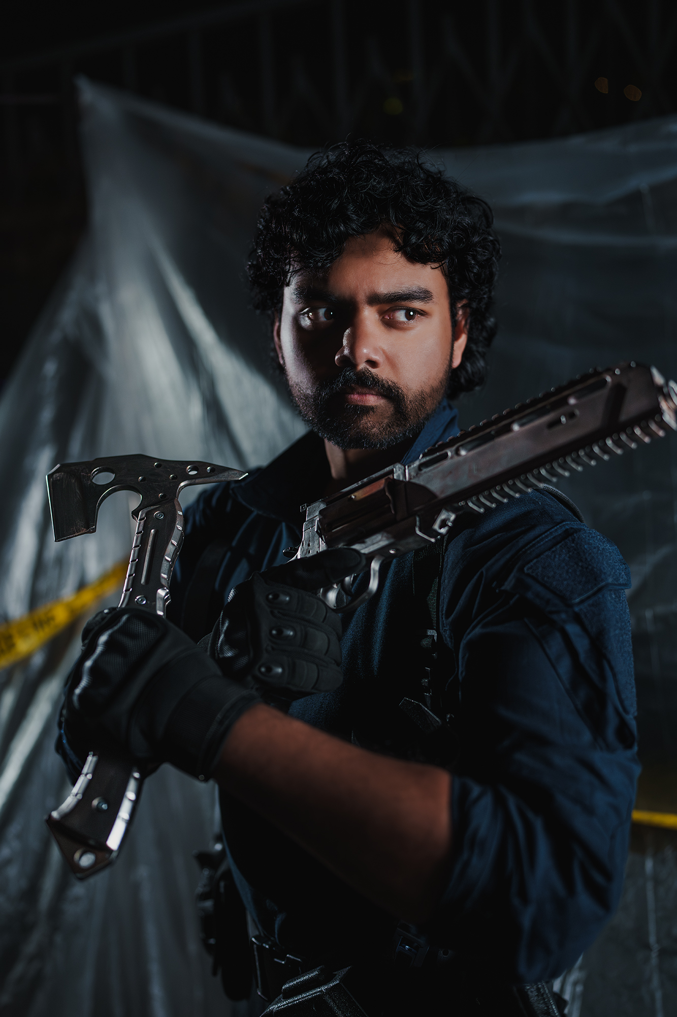 Man with curly hair and beard holding a large weapon, wearing dark clothing, in a dark setting with a tarp background.