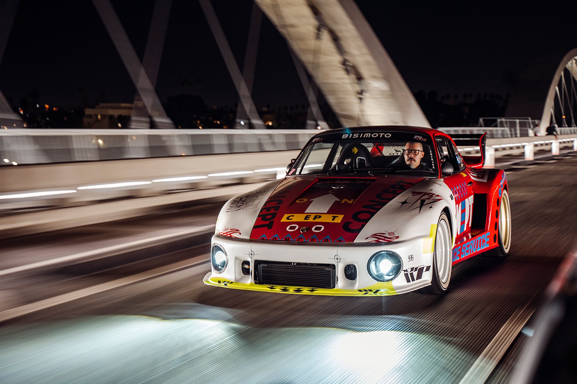 Race car on bridge at night with driver visible, headlights on, and city lights in background.