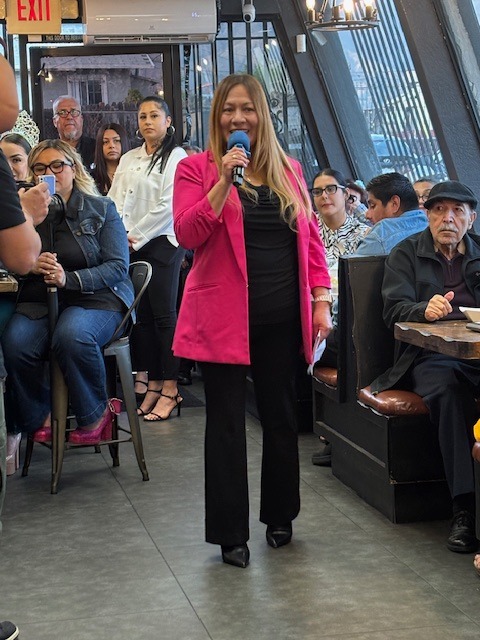 Woman in pink blazer speaking into microphone in a restaurant with seated people around her.