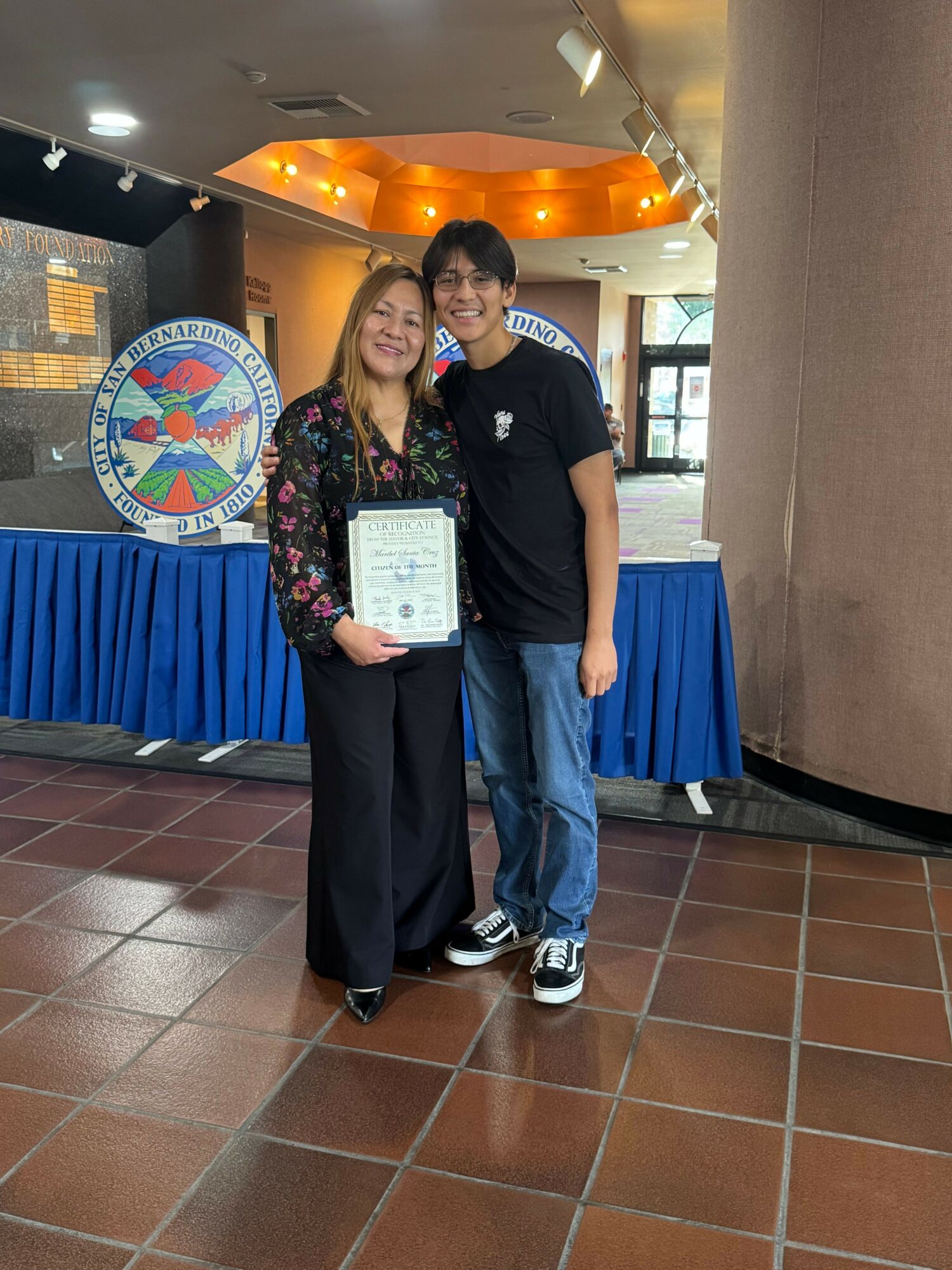 Woman holding a certificate, standing indoors near a blue table and a circular logo, with a window in the background.