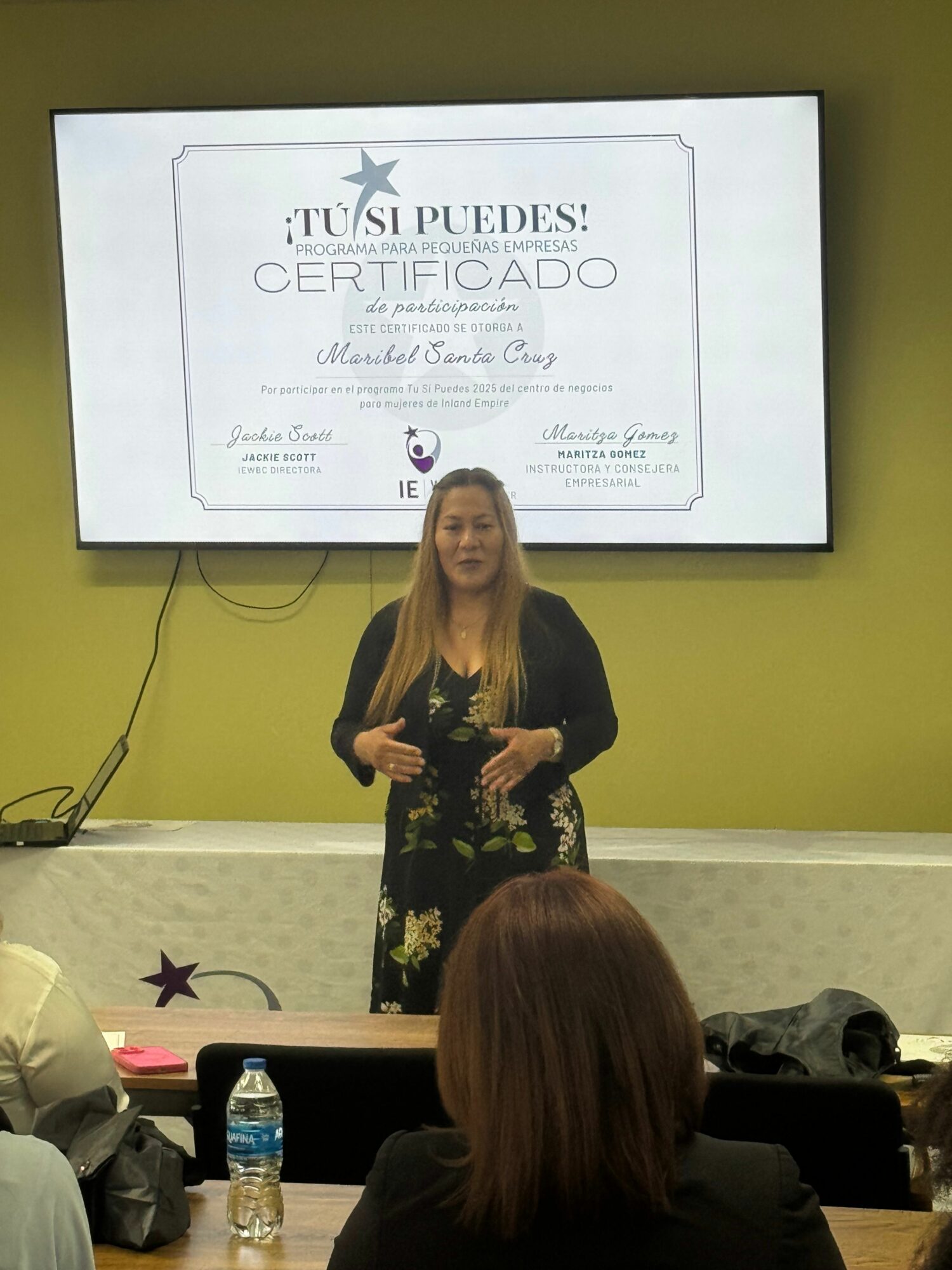 Woman standing and speaking in front of a screen displaying a certificate, with people seated in the foreground.