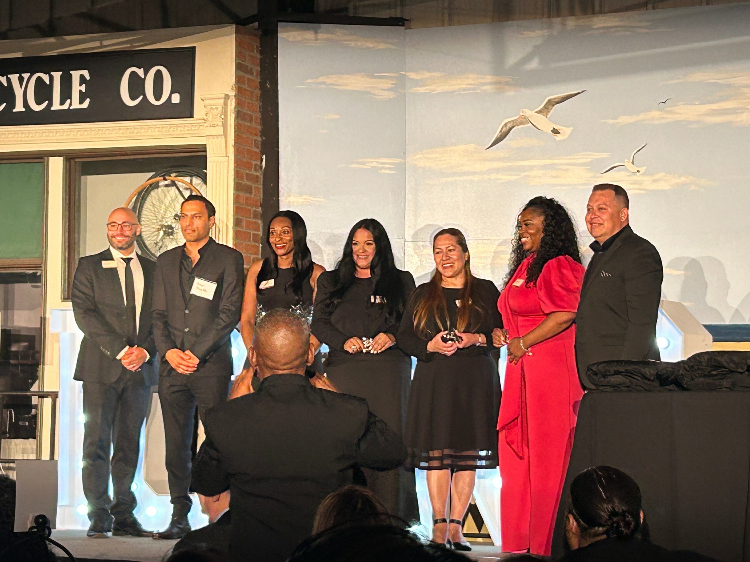Six people stand on stage in front of a painted backdrop with flying birds, posing for a photo, with a man in the foreground taking the picture.