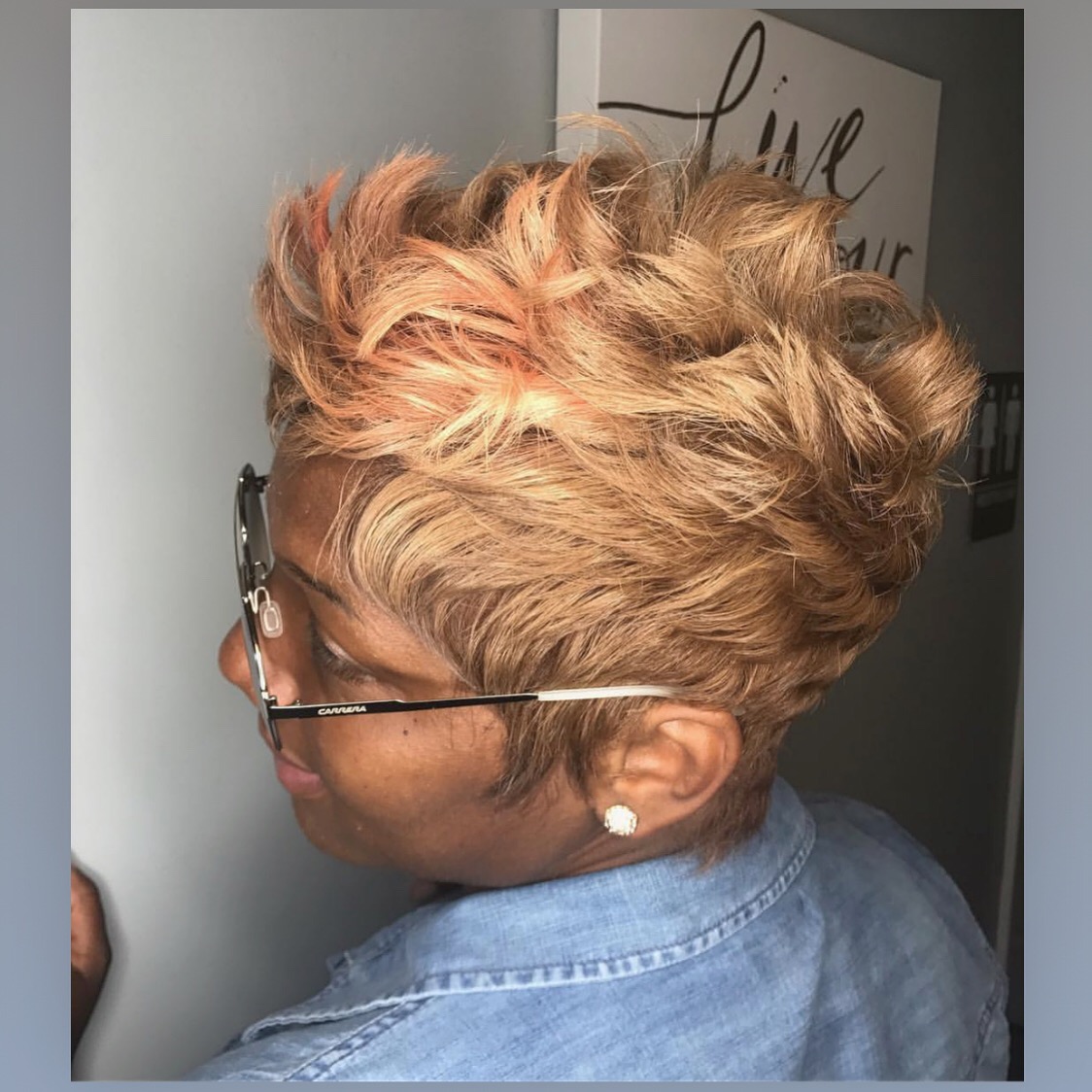 Side view of a person with short, curly, blonde and pink hair, wearing glasses and a denim shirt, indoors.