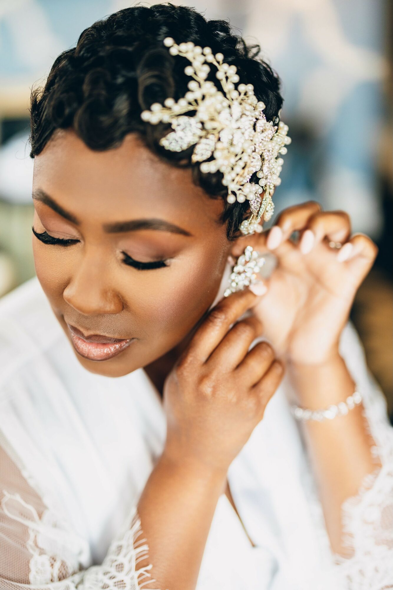 Close-up of a woman with short curly hair, wearing a decorative hairpiece, adjusting an earring, with a soft background.