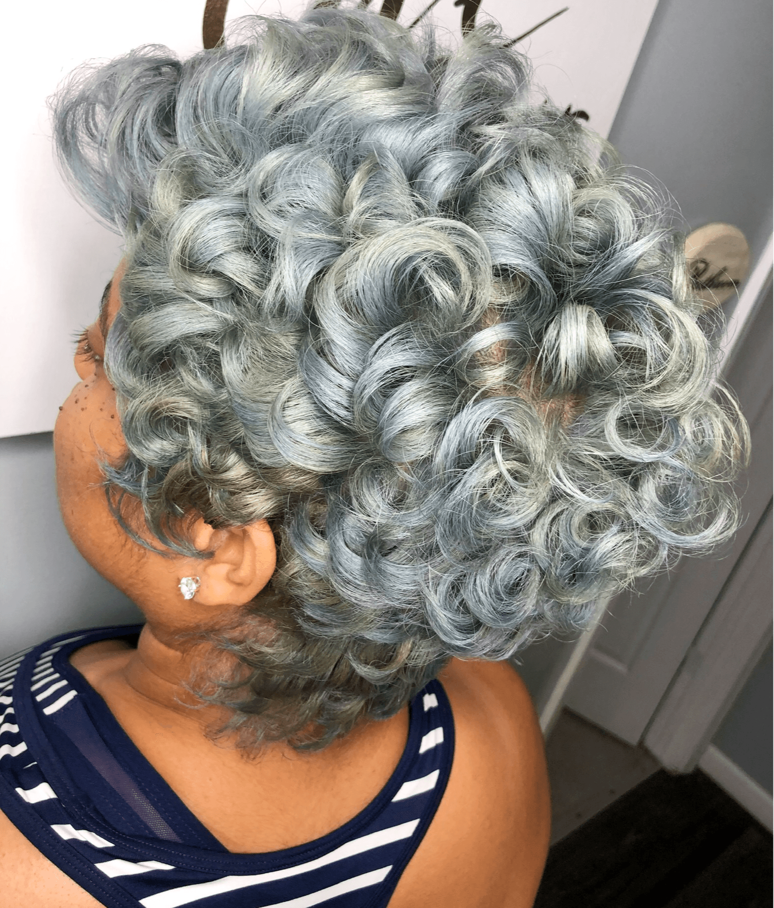 Person with short, curly, silver-gray hair styled in tight ringlets, wearing a striped top, seen from behind.