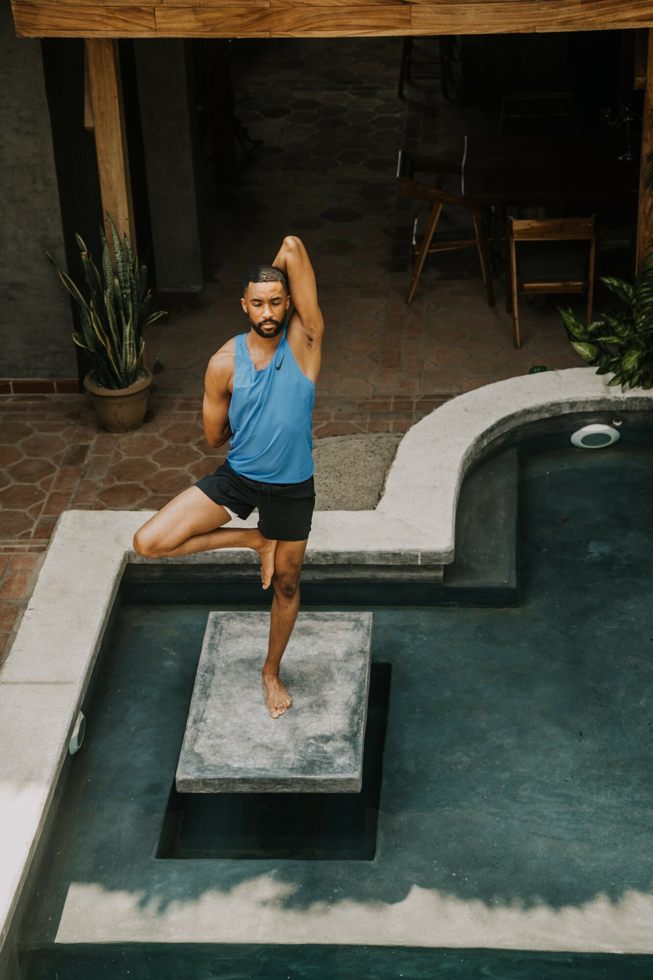 Person standing on a raised platform in a pool, stretching arm behind head, wearing a blue tank top and black shorts.