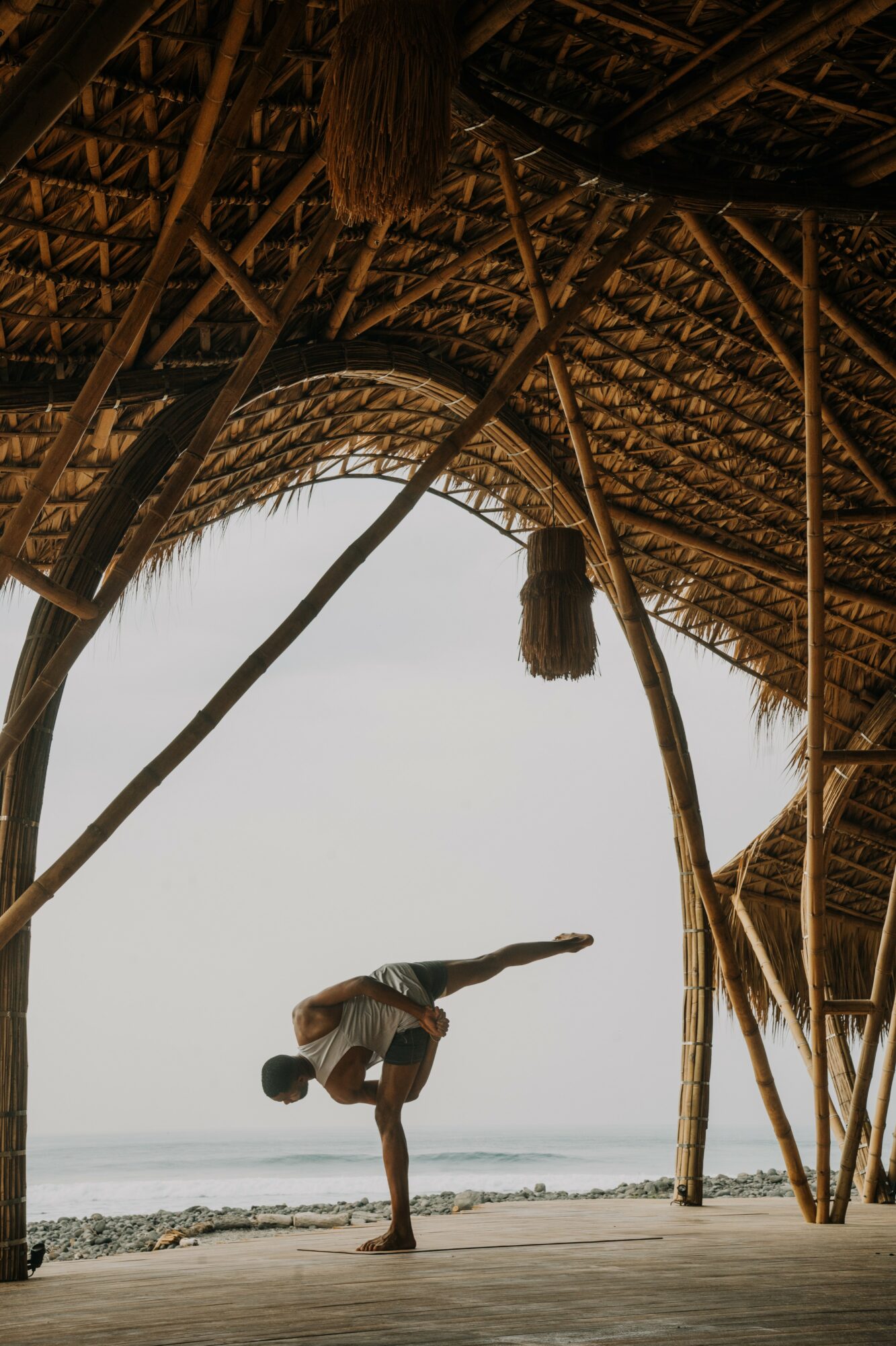 Person performing a handstand on a beach under a thatched roof structure, with the ocean in the background.