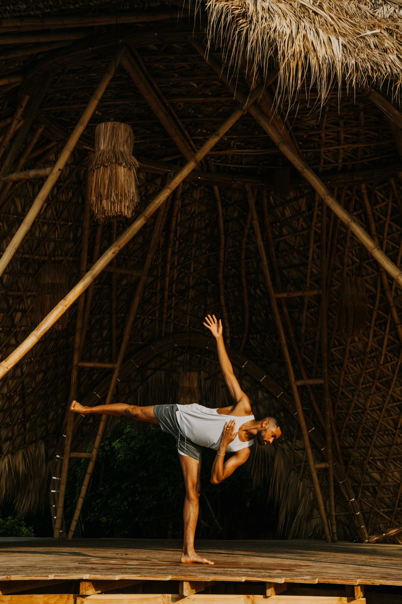 Person performing a dance or yoga pose on a wooden stage with a thatched roof structure in the background.