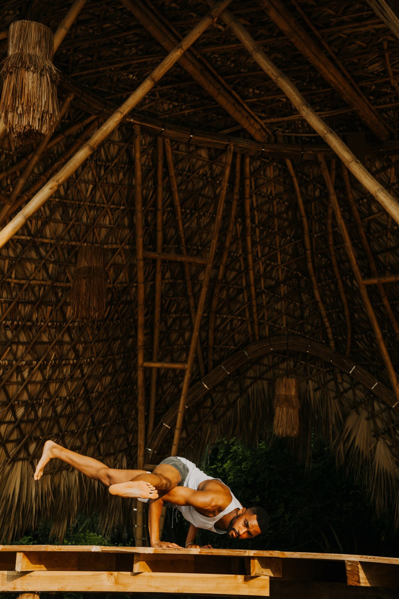 Person performing a yoga pose on a wooden platform inside a thatched structure, with a curved entrance and natural materials.