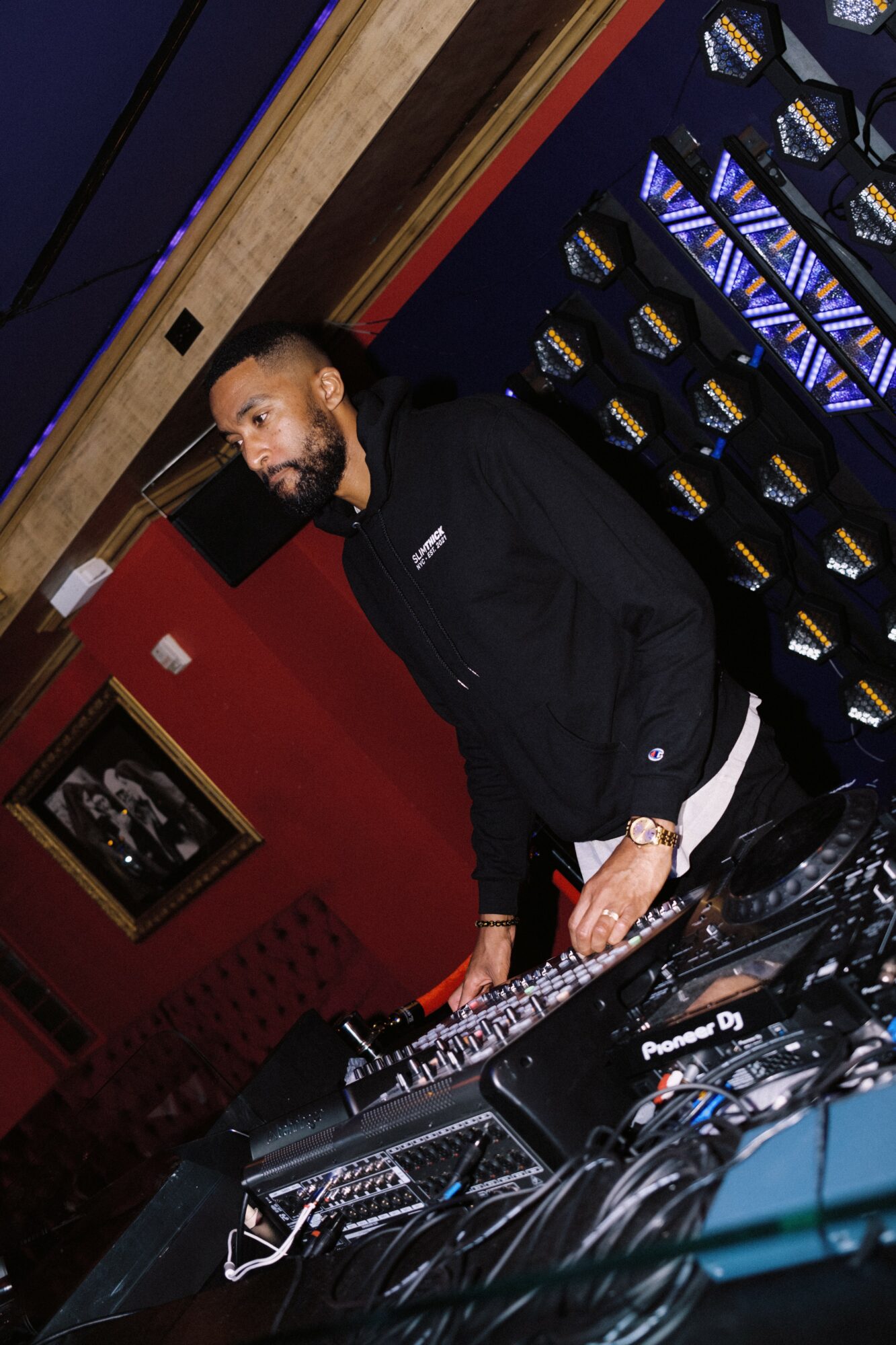 Man with beard in black hoodie standing behind DJ equipment in a dimly lit room with red and blue walls.