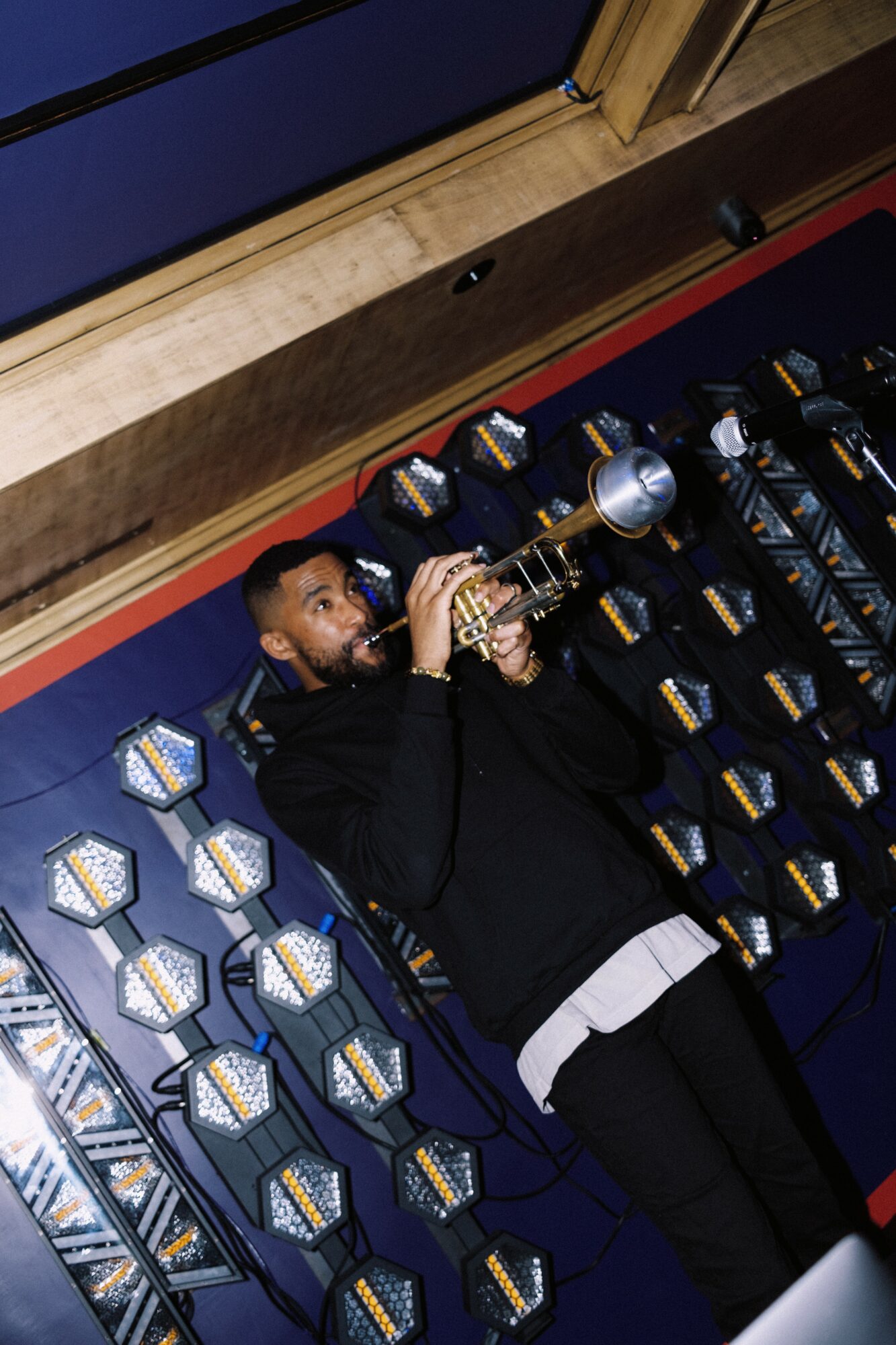 Person lying on floor surrounded by stacked boxes, playing a trumpet, with a wooden ceiling above.