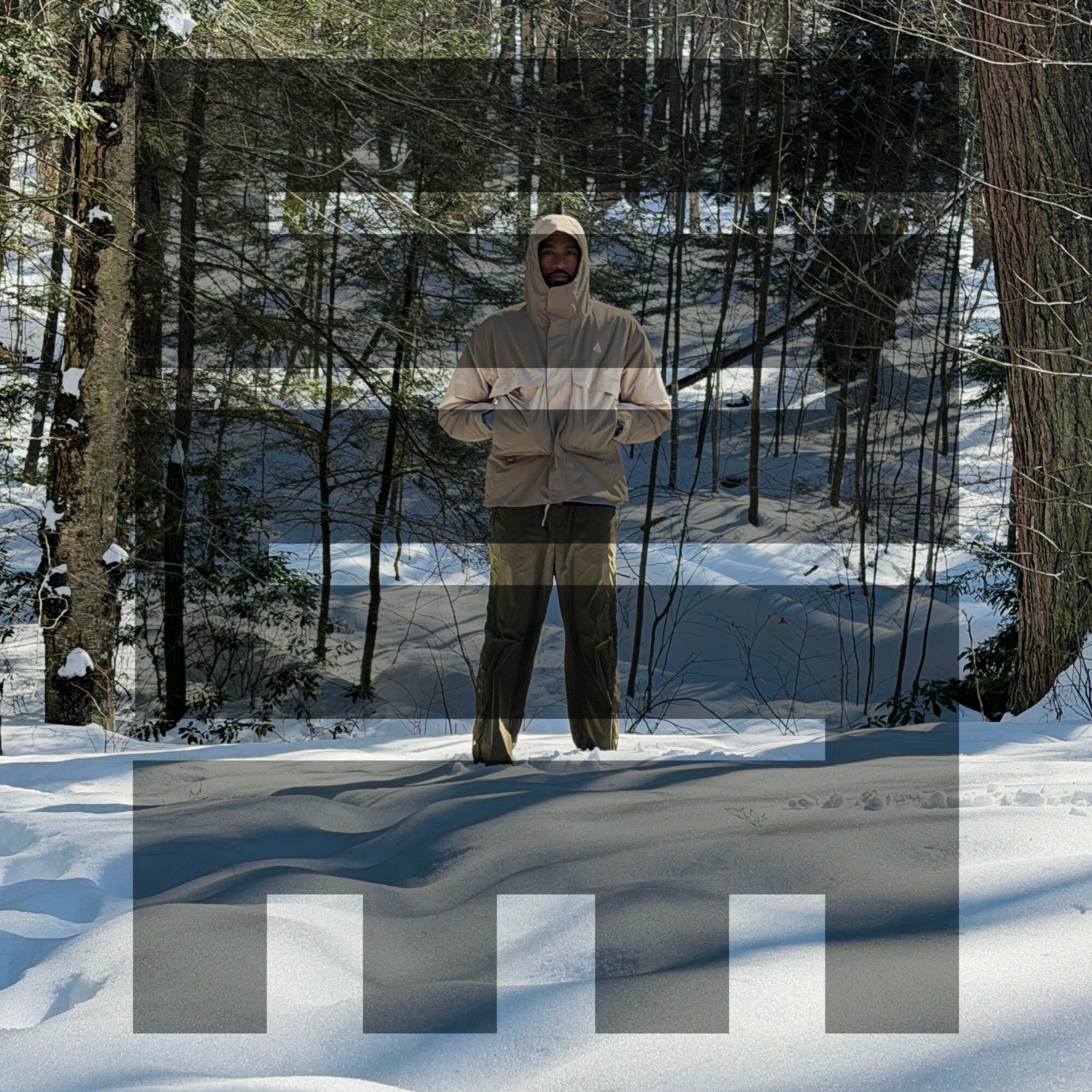 Person standing in snowy forest wearing a hooded jacket and pants, surrounded by trees and snow, with a reflective surface behind.