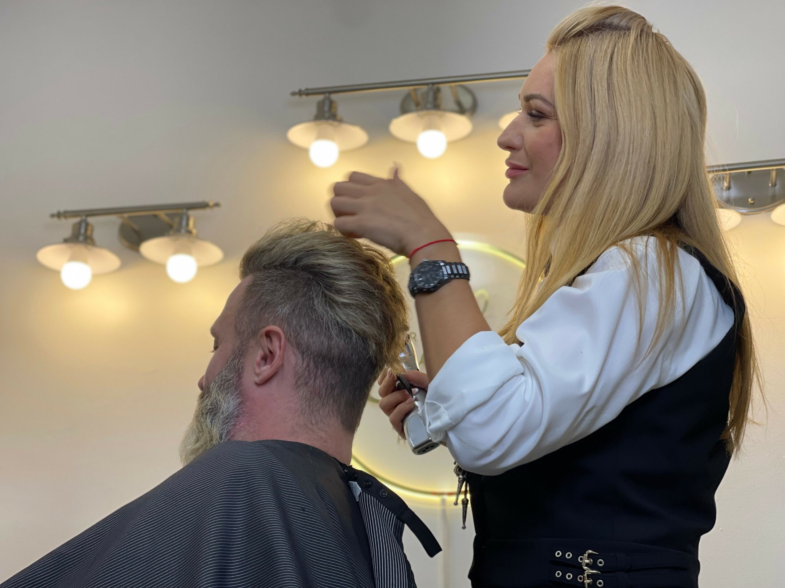 A woman with long blonde hair styles a man's hair in a salon, with bright lights overhead.