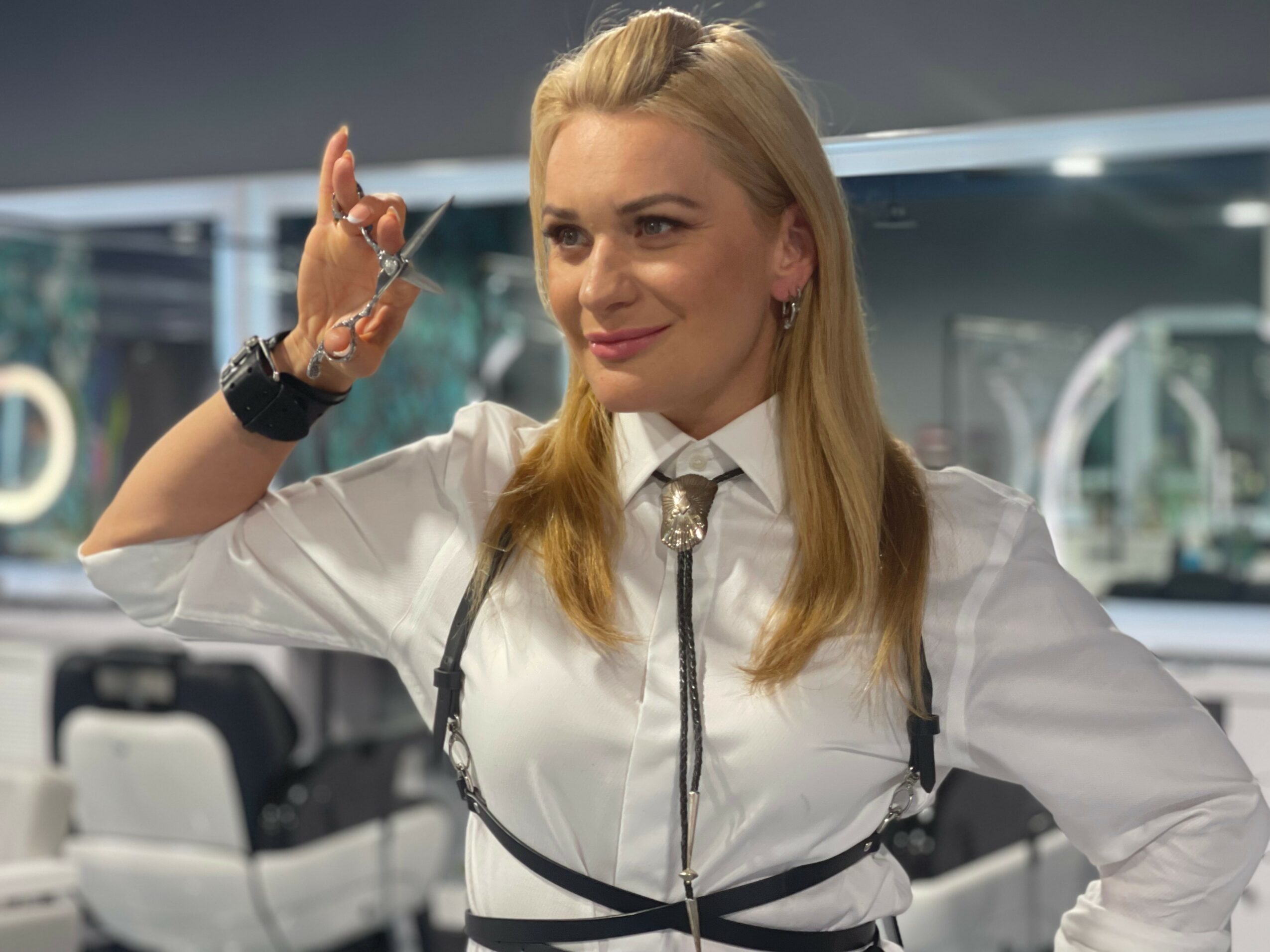 Woman with blonde hair smiling, wearing a white shirt and accessories, in a modern indoor setting.