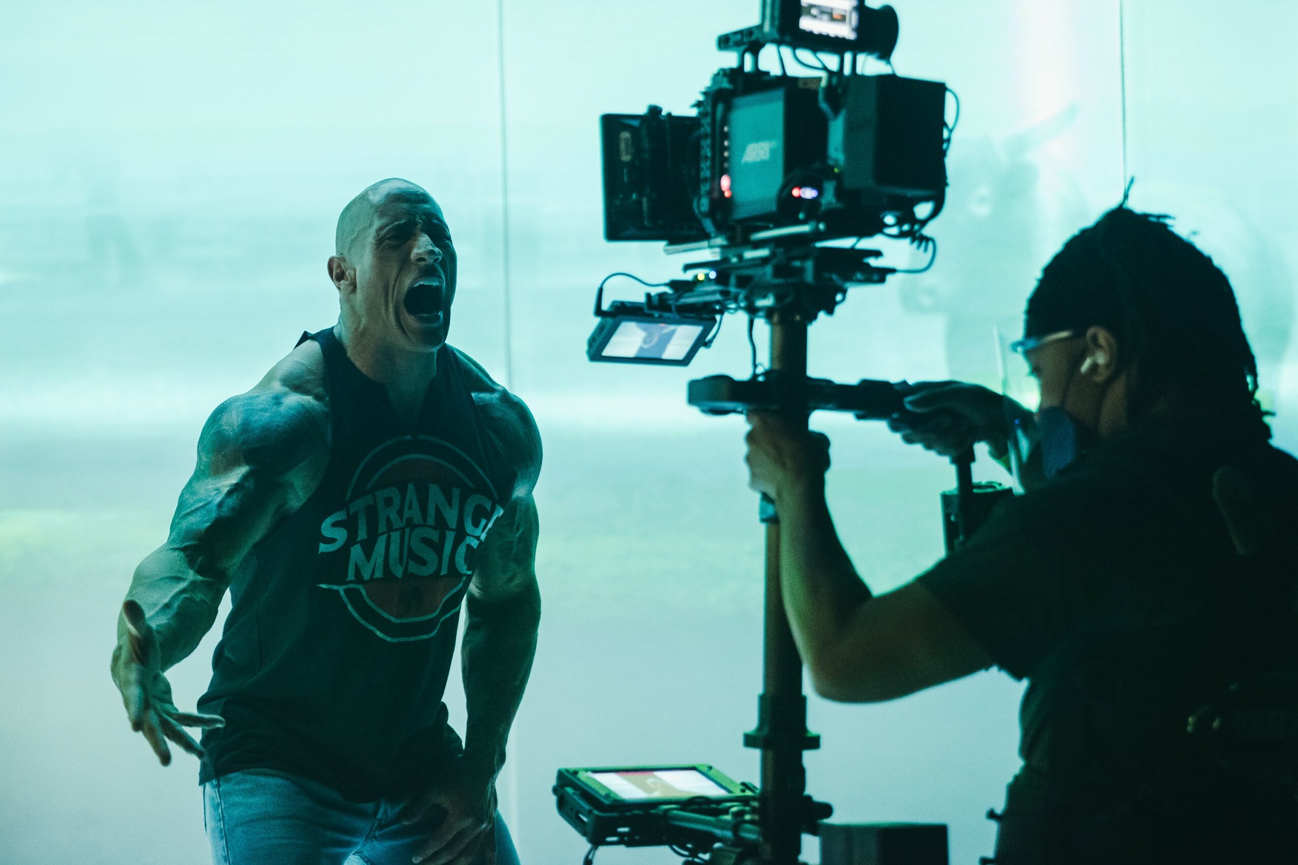 Person singing passionately during a performance, with a camera operator filming in a bright setting.